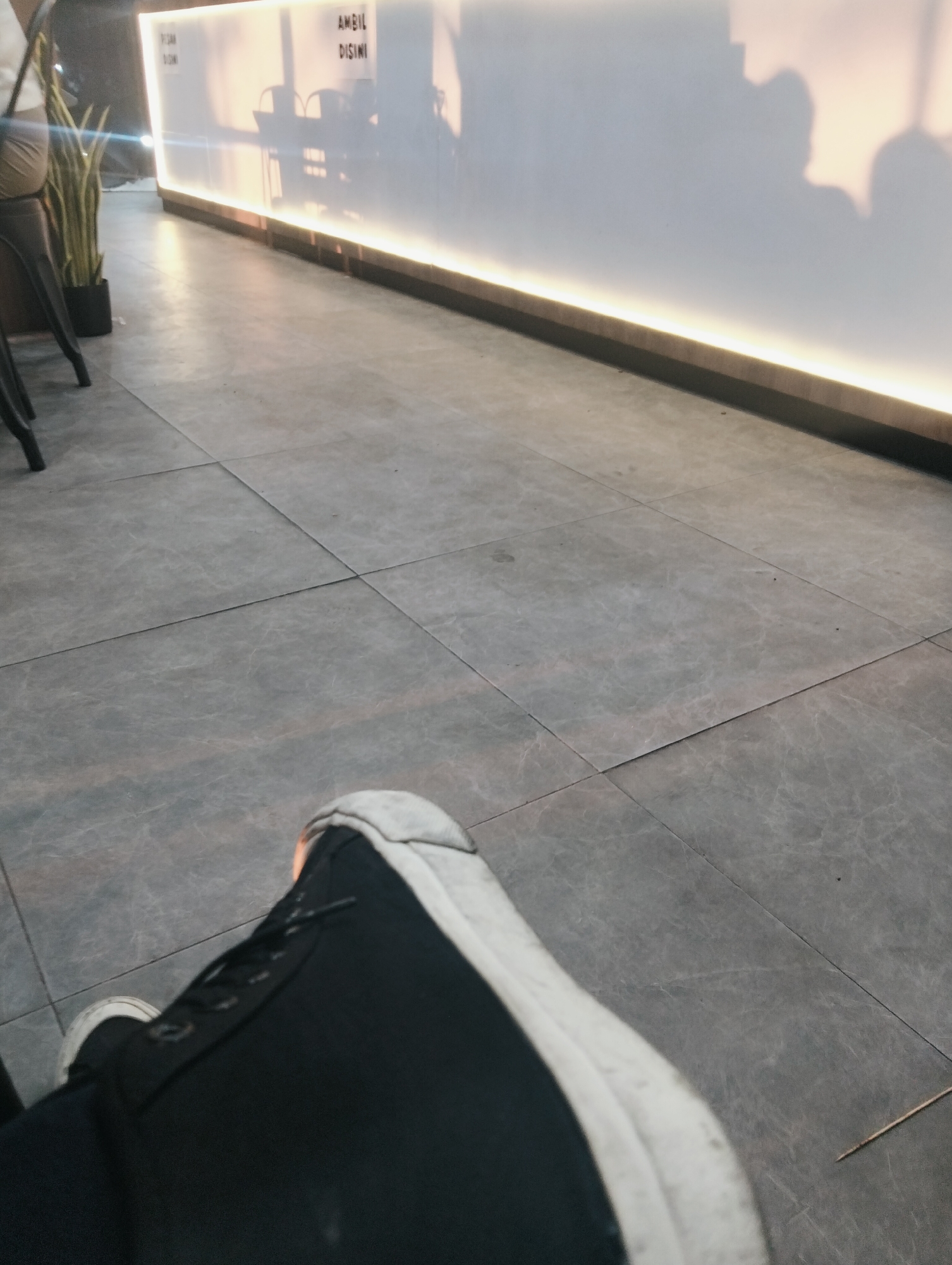 The image shows a close-up view of a person's feet wearing black sneakers with white soles, positioned on a tiled floor. In the background, a brightly lit counter or wall is visible, with the words "AMBIL DI SINI" (Take Here in Indonesian) displayed. Shadows of people are cast on the wall. A potted plant with long, green leaves is seen to the left of the counter. The overall setting appears to be an indoor public space, potentially a cafe or restaurant, in the city of Medan, Indonesia. The lighting is soft and warm, with a prominent strip of light illuminating the base of the counter.