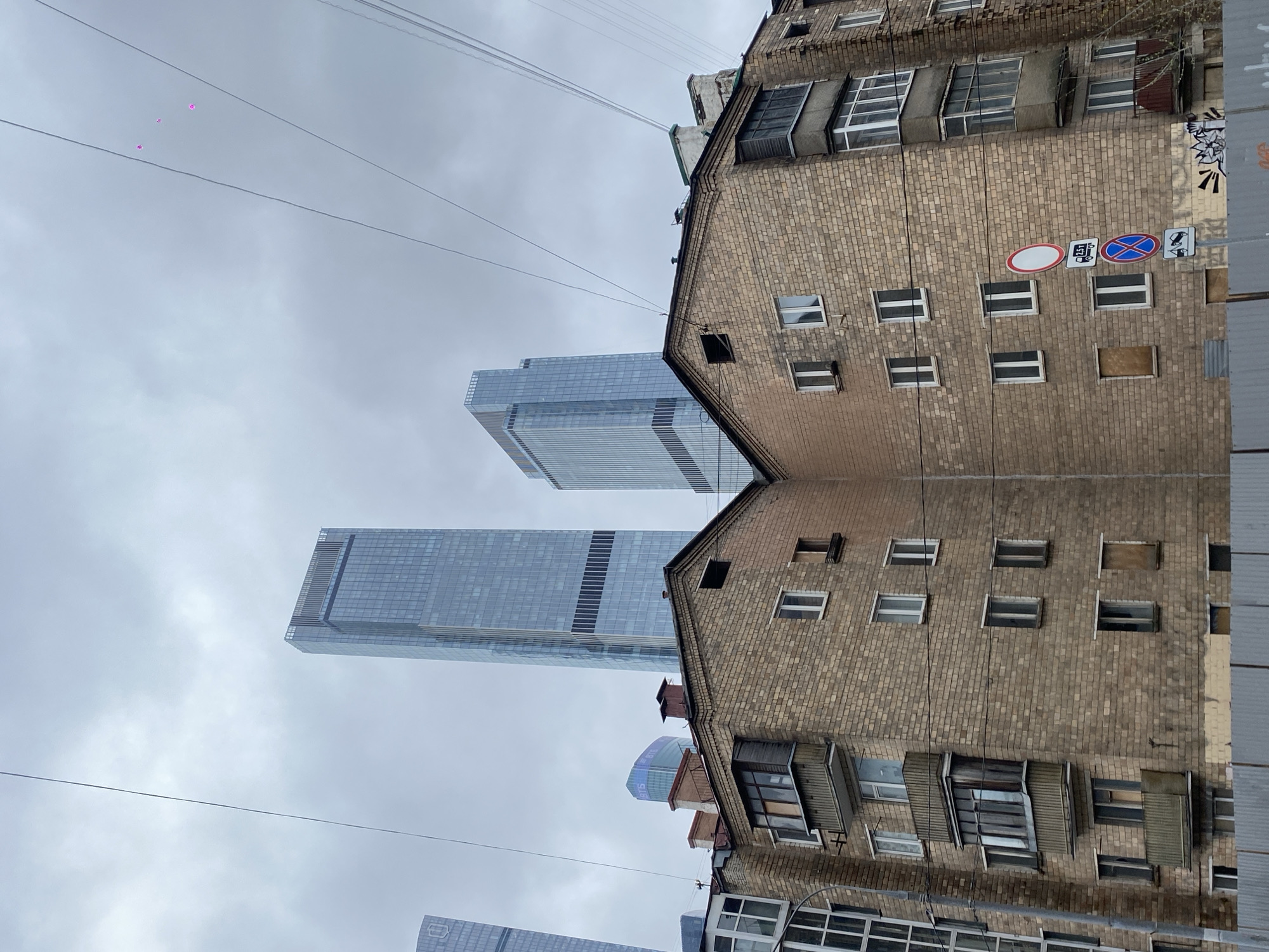 The image depicts a street scene in Khimki, Russia. It shows a juxtaposition of old and new architecture. A brick apartment building stands on the right side, displaying multiple windows and some balconies. Various street signs are visible on the wall. To the left, the modern glass facade of the Khimki Arena towers in the background, partially obscured by overcast skies. A network of electrical wires stretches across the scene. The buildings appear to be mirrored.