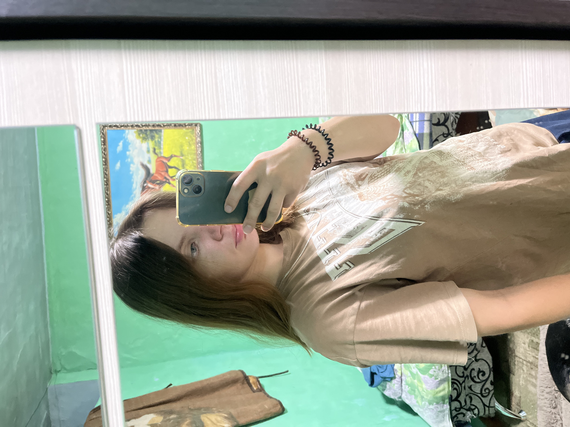 In a room with green walls, a young woman takes a selfie in a mirror. She is wearing a light brown t-shirt and dark blue pants. Her hair is long and brown. She is holding a dark-colored smartphone in her right hand. On her left wrist, she wears two dark-colored spiral hair ties, serving as bracelets. In the mirror's reflection, a framed picture of two horses in a field is visible on the wall behind her. To the left, a brown garment hangs on the wall. The room appears to be a bedroom, with a bed visible in the background, covered with blankets. The overall impression is of a casual, personal moment captured in a private space.
