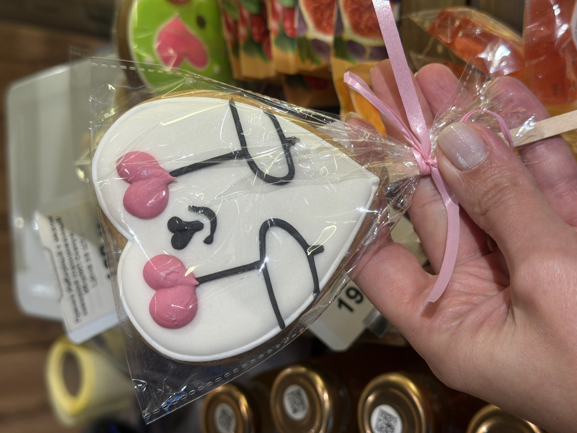 A hand holds a heart-shaped cookie, decorated with white icing and a drawing resembling a cow, wrapped in clear plastic and tied with a pink ribbon. The background includes other packaged treats, jars, and a tag with the number "19" visible. The location is Rostov-on-Don, Russia, as indicated by the presence of Cyrillic text on some of the packaging.