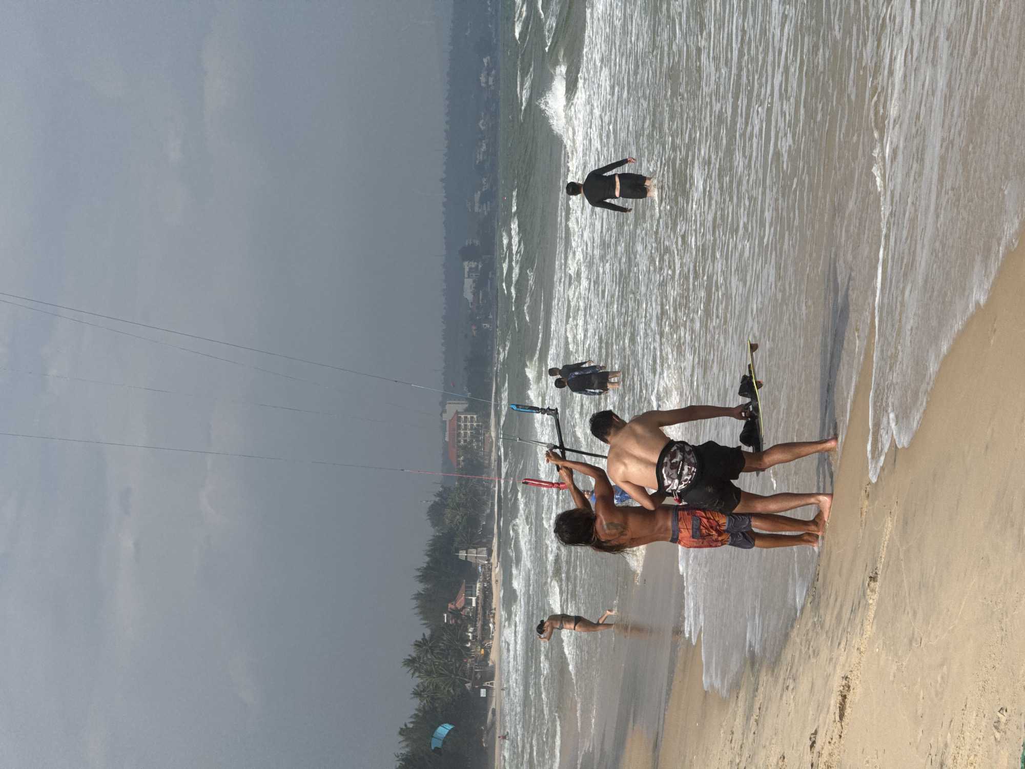 At a beach in Mũi Né, Vietnam, two men are preparing to kite surf. One man, wearing orange and grey patterned shorts, is facing away from the camera and has his back to the water. The other man, wearing black shorts and a black harness, is standing in the shallow water facing the first man. He is holding a kite bar and is in the process of teaching or demonstrating. Further out in the water, three other individuals are visible. Two are standing in the water, one with their back to the camera wearing a black wetsuit, and another facing left. A third person is further out in the water. The beach itself is sandy with gentle waves breaking on the shore. In the background, a line of palm trees and buildings is visible, with a cloudy, overcast sky overhead. A blue kite is visible in the distance to the left.
