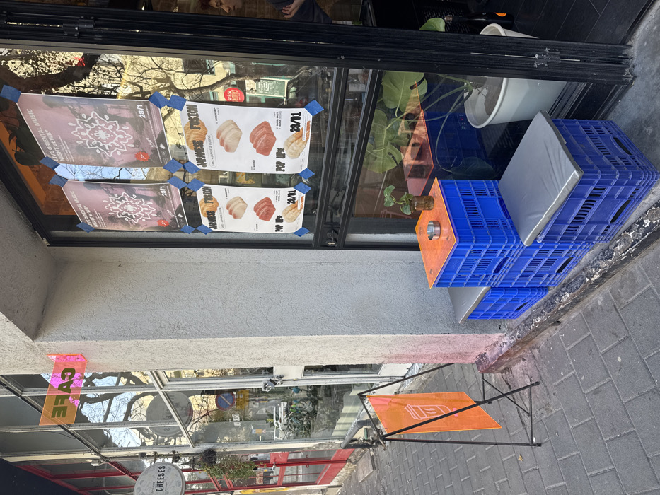 A store window has posters taped to it advertising "Live Oriental Music" on the 28th and a "Japanese Fusion" pop-up on November 24th. Outside the store is a seating area with blue plastic crates stacked to form a low table with gray cushions. A small table with an orange top sits nearby. A sign reading "Cafe" hangs in the window, and another sign reads "Cheeses". The image captures a street scene with reflections in the window.