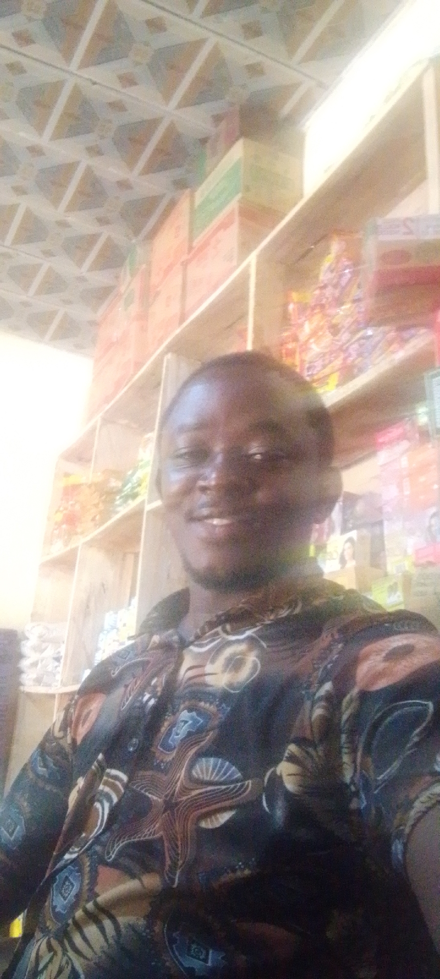 The image features a man, appearing to be of African descent, smiling and posing for a close-up photo. He has short dark hair and a neatly trimmed beard, and is wearing a dark, short-sleeved button-up shirt with an intricate pattern of starfish, shells, and abstract designs in shades of brown, blue, and white.
Behind him is the interior of what appears to be a small shop or market stall, consistent with a location like Adankolo, Nigeria. Wooden shelves are extensively stocked with various products. These include stacked boxes of different colors (red, green, yellow), as well as numerous packaged goods, some appearing to be snacks or confectionery in clear plastic bags with colorful contents. On a lower shelf to the right, packages featuring images of women suggest products like cosmetics or hair care items. The ceiling visible at the top of the frame has a distinctive geometric pattern of interlocking triangles and squares.
The man's direct gaze and broad smile convey a friendly and positive emotion. The lighting is bright, indicating an indoor daytime setting. No specific text is clearly legible on any of the products or packaging due to the image resolution. The scene effectively captures the atmosphere of a local, well-stocked retail environment.