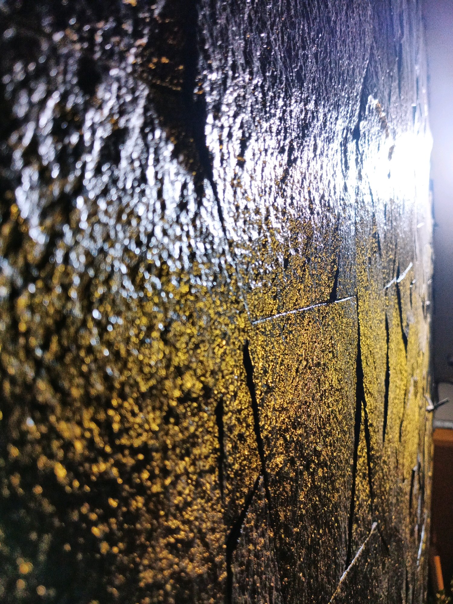 This is a close-up, low-angle shot of a textured, dark wall that appears to be made of stone or concrete. The wall is partially illuminated by a bright light source from the right, casting highlights and creating a shimmering effect across its surface. The texture of the wall is rough and uneven, with visible grooves and indentations that create patterns of light and shadow. There are also small, irregular specks of a golden or yellow material adhered to the wall, which catch the light and add to the shimmering appearance. Some dark, vertical lines, possibly cracks or painted strokes, are also visible on the surface. The foreground is out of focus, showing a bokeh effect of blurred circles of light and dark. The background on the right is dimmer, suggesting an indoor environment or a shadowed outdoor area, with a hint of a vertical structure. The image does not contain any people, animals, or discernible objects from the city of Bandung, Indonesia. It is an abstract capture of a textured wall surface.
