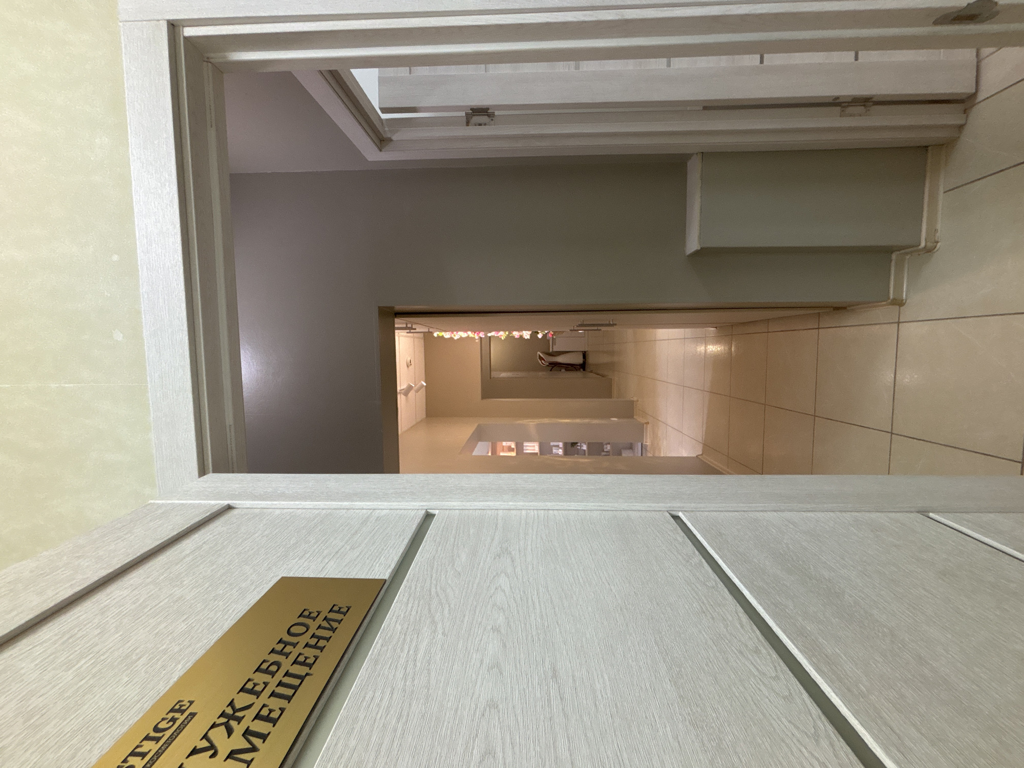 The image shows a slightly ajar door, angled upwards to reveal a hallway or waiting area beyond. The door itself is light-colored with vertical recessed panels. A gold-colored sign with lettering is attached to the door, featuring text that includes the words "PRESTIGE" and lettering in Cyrillic. The hallway has a beige-tiled floor and light-colored walls. In the distance are shelves and what appears to be a waiting area with seating. Based on the text on the sign, and given the context of your prompt, this may be located in Vsevolozhsk, Russia.