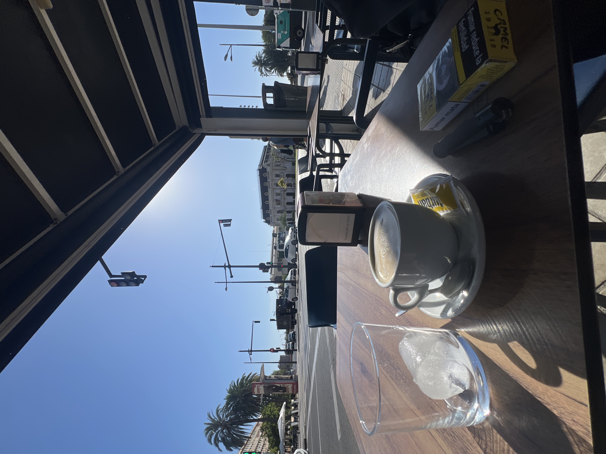 The image depicts a sunlit outdoor cafe scene with a table in the foreground. On the wooden table, there is a partially filled glass of ice, a white ceramic cup of coffee on a saucer with a spoon, a packet of Camel cigarettes, and a black vape pen. In the background, a street is visible with traffic, palm trees, and buildings. The scene appears to be a street-level cafe in Valencia, Spain, with architectural elements like awnings and outdoor seating visible. The sunlight casts strong shadows on the table, indicating a bright, sunny day. The sky is clear and blue, and there are traffic lights and street lamps.