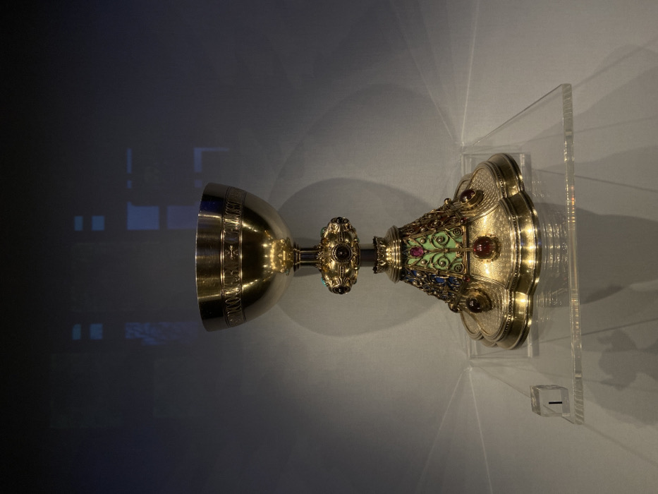 A highly ornate gilded chalice is presented centrally on a clear acrylic display shelf, indicative of a museum or gallery exhibition space in London, United Kingdom. The chalice is constructed from gold-colored metal, featuring a polished bowl engraved with a band of text and small crosses below its rim. A central, spherical knop on the stem is studded with multiple oval and round gemstones in red, purple, and turquoise. The multi-lobed base is elaborately decorated with segments of green enamel featuring gold filigree, interspersed with triangular areas containing blue and red gemstones, and several larger, round red-purple gemstones encircling the base. The top surface of the base displays fine patterned engraving. A clear cubic marker block, inscribed with the numeral '1', is positioned on the shelf to the left of the chalice. The background is a dark, reflective surface, displaying indistinct luminous rectangular reflections. No human subjects or discernible actions are depicted.