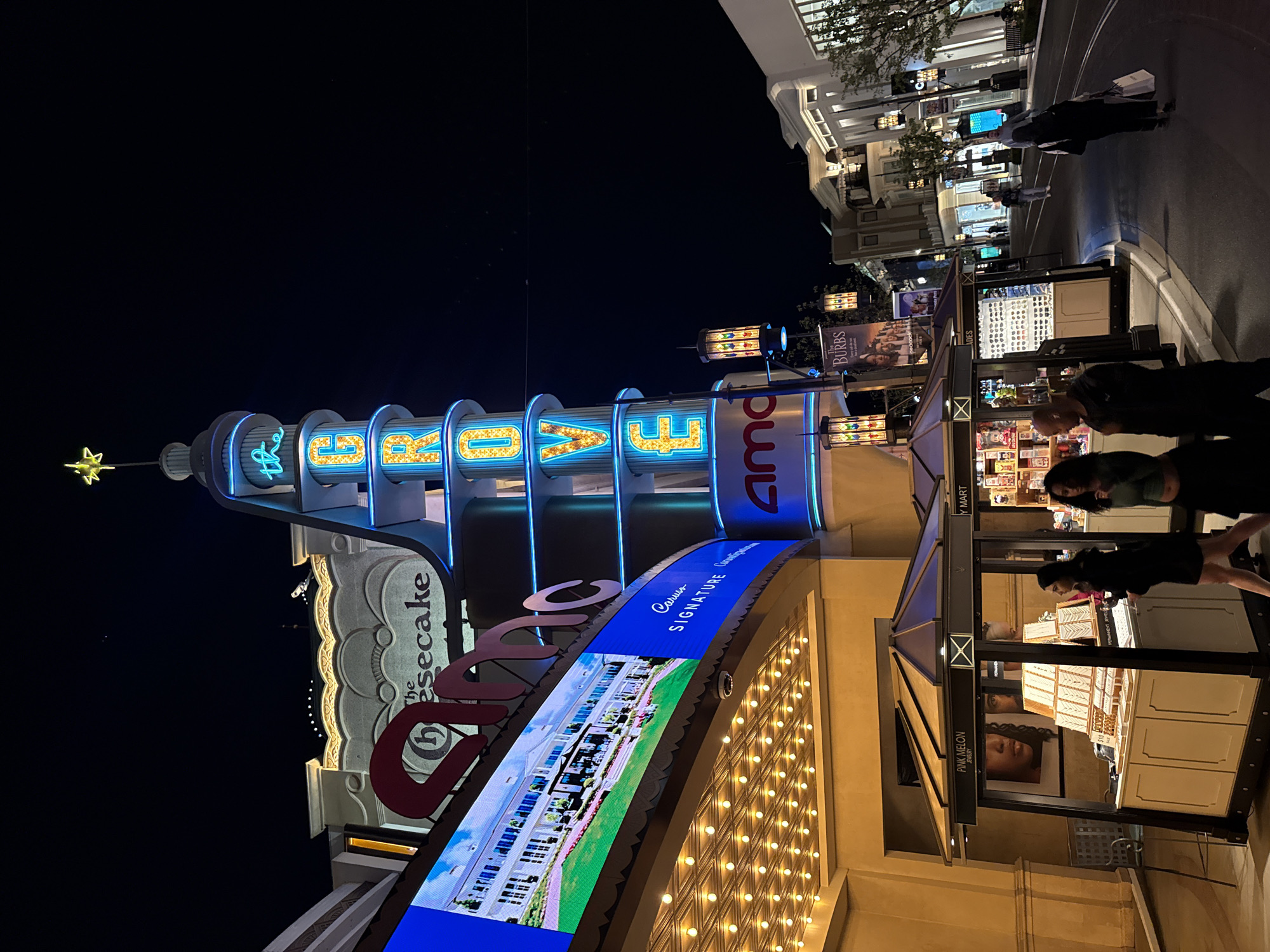 Here's a concise description of the scene:
The image captures an outdoor night scene in Los Angeles, likely at The Grove shopping center. Notable landmarks include "The Grove" tower with illuminated signage, an AMC movie theater, and The Cheesecake Factory restaurant. The scene is brightly lit, featuring people walking past shops and kiosks. A stand selling Pink Melon jewelry is visible. Overall, it presents a bustling urban ambiance at night.
