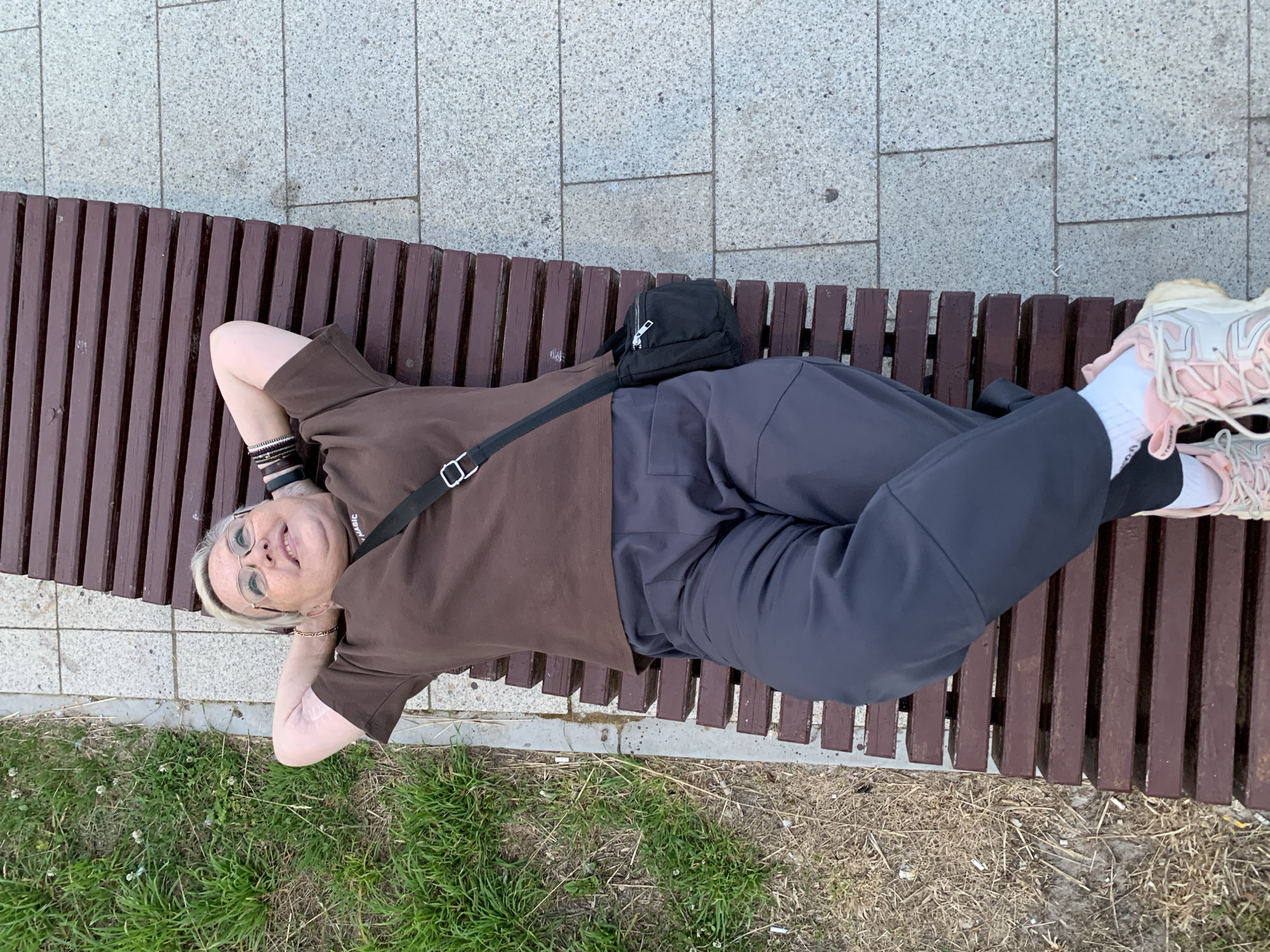 A blonde woman with glasses is lying on a wooden park bench, her hands behind her head, looking up and smiling. She is wearing a brown t-shirt, dark gray pants, and pink and white sneakers. A black crossbody bag is hanging over her shoulder. The bench is situated on a paved walkway next to a grassy area. The location is the Chaikovsky city park in Chaikovsky, Russia.