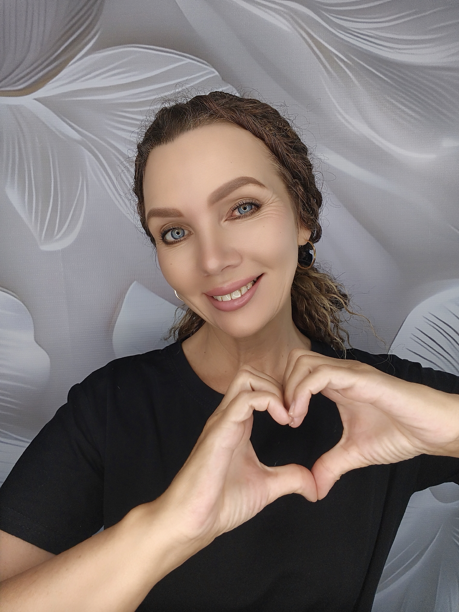 A woman with blue eyes and curly brown hair is shown from the chest up, making a heart shape with her hands. She is wearing a black t-shirt and large hoop earrings. The background is a light-colored fabric with a subtle floral pattern.