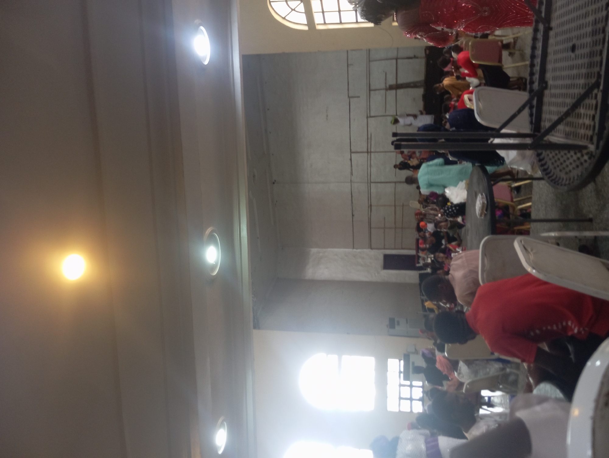 The image shows the interior of a large room, possibly a church or event hall, in Onitsha, Nigeria. Rows of seated people fill the space. Most of the people are facing towards what appears to be the front of the room. White plastic chairs and metal tables are visible. The room has a high ceiling with inset lights. There are arched windows at both ends of the room. The walls are painted white. The lighting is bright.