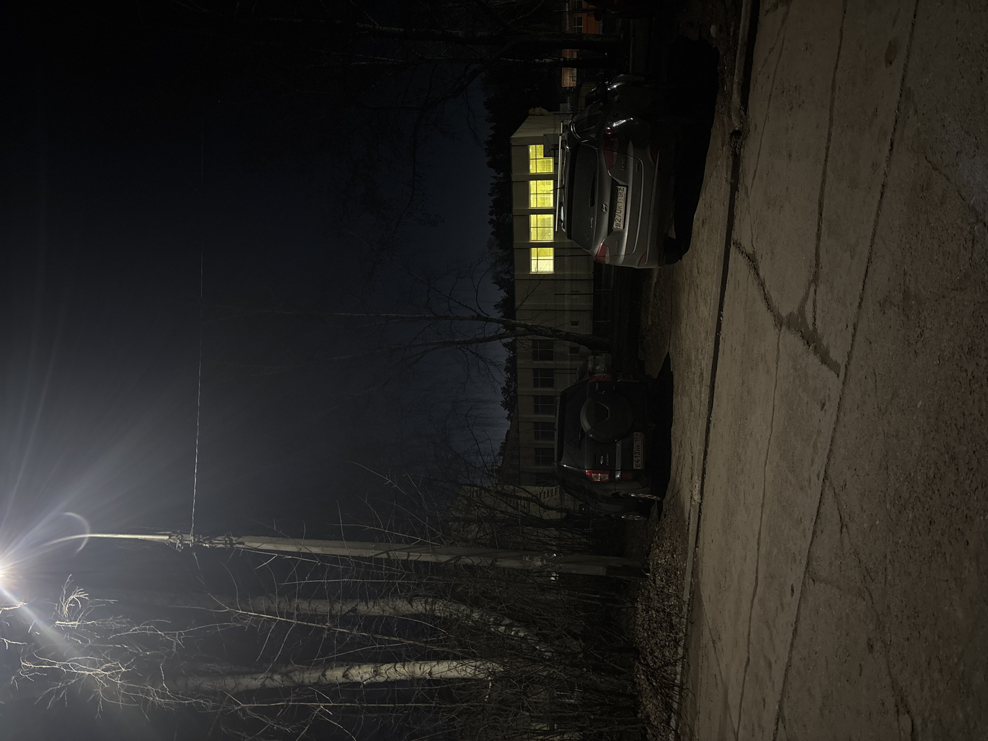 The image depicts a nighttime scene in an urban or suburban environment, likely in Chaikovsky, Russia, given the location context. The perspective is from ground level, looking towards a building and parked cars under a dark sky.
A bright streetlight on the left side of the frame casts a strong light, creating lens flare and illuminating the bare, skeletal branches of nearby trees in the foreground. The ground consists of concrete pavement or a sidewalk, showing various cracks and lines.
Two cars are visible. A silver Hyundai sedan is parked on the right, partially on the pavement. Its rear license plate, "B201KT 152", is clearly discernible, indicating a Russian registration. Behind it, a dark SUV, possibly a Chery (text "CHERY" is visible on the spare tire cover), is also parked. Its license plate, "E413HK 152", is likewise visible.
In the mid-ground, behind the vehicles and trees, stands a multi-story building. Several of its windows glow with a bright, warm yellow light, contrasting with the dark surroundings. The outlines of a darker forest or dense tree line are visible behind the building.
The scene is quiet and devoid of people, characterized by the artificial illumination piercing the night. The bare trees suggest the season is late autumn or winter.