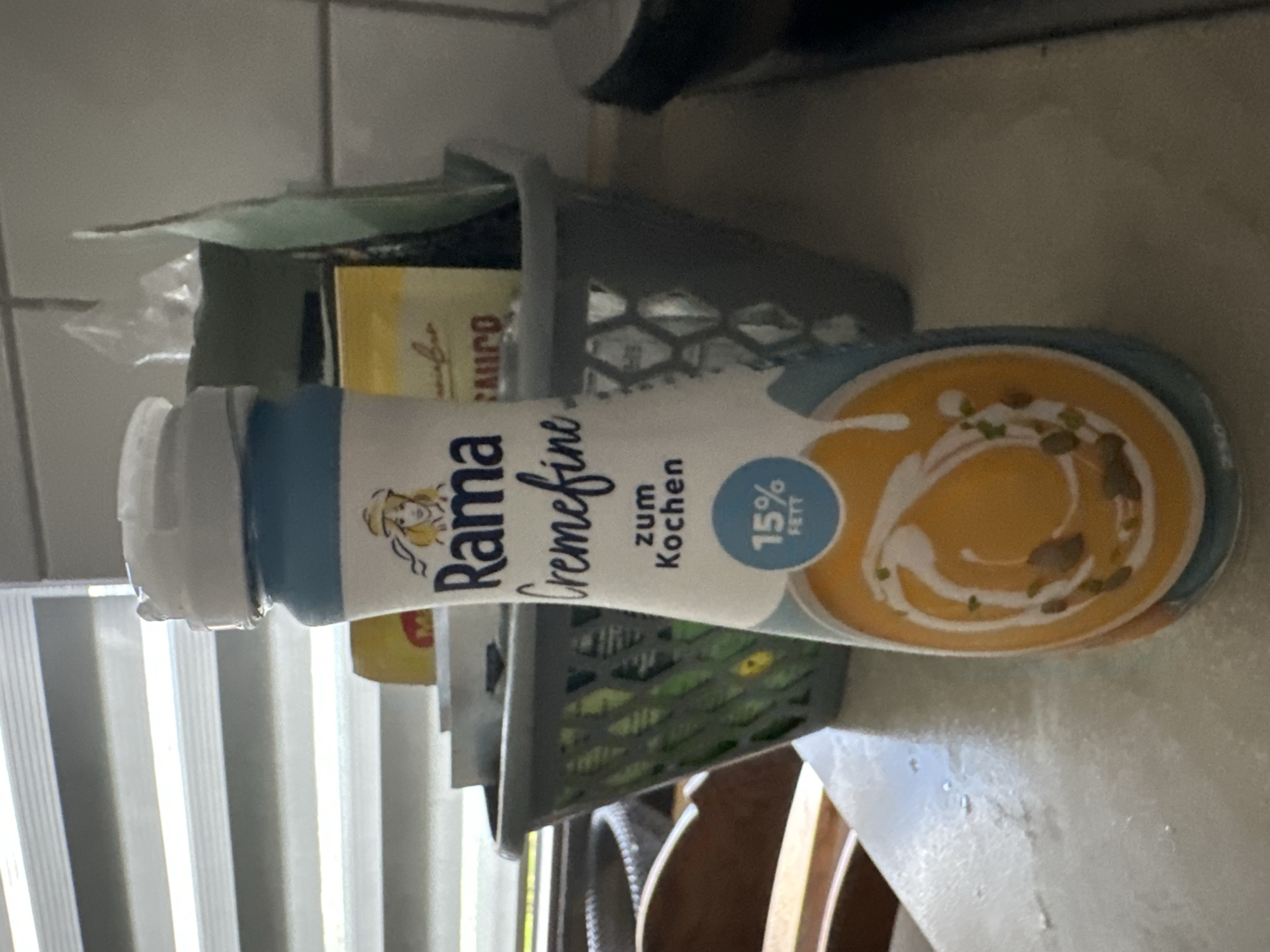 The image shows a bottle of Rama Cremefine 15% Fett zum Kochen, a cooking cream product. The bottle is white with blue and yellow labeling. The label features the Rama logo, which includes a woman with blonde hair wearing a hat, and an illustration of a bowl of soup topped with cream and seeds. Text on the bottle reads "Rama Cremefine zum Kochen" and "15% Fett". The bottle is placed on a countertop in what appears to be a kitchen or pantry area. In the background, a grey plastic basket holds some items, and a stack of blinds is visible on the left. The lighting suggests it is daytime, and the setting is likely indoors, possibly in Krefeld, Germany, given the context. There are no people visible in the image. The overall impression is that of a product shot or a casual photograph taken in a home environment.