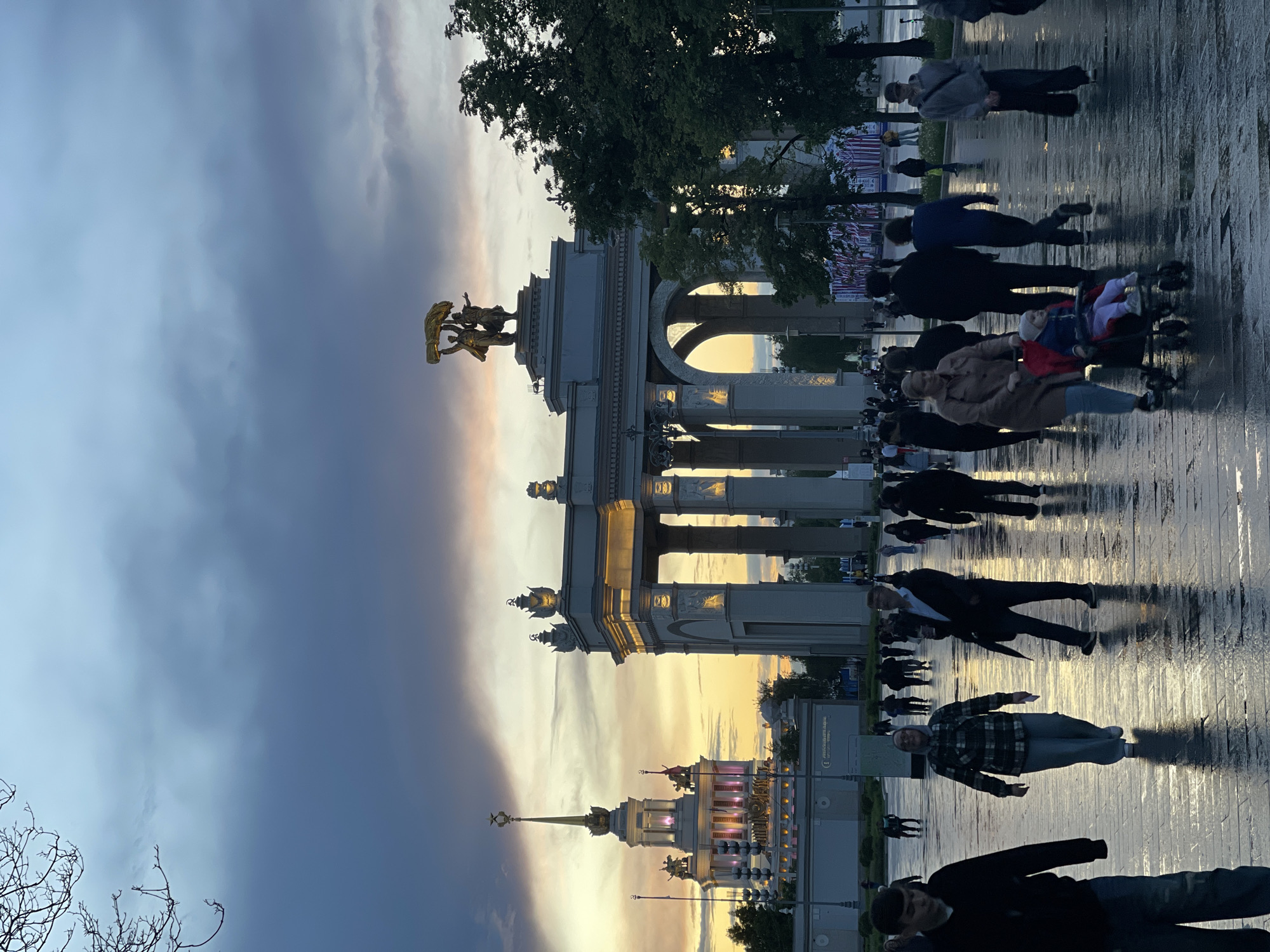The image depicts a twilight scene at the All-Russia Exhibition Center (VDNKh) in Moscow, Russia. A large, ornate archway with a golden statue on top stands prominently in the frame, reflecting the yellow-orange light of the sunset. Below the arch, the ground appears wet and reflective, possibly due to recent rain. Pedestrians are walking around the scene, some of whom are pushing strollers. In the background, the illuminated spire of another building within the VDNKh complex is visible. The sky above is a mix of dark storm clouds and the fading light of the sunset.