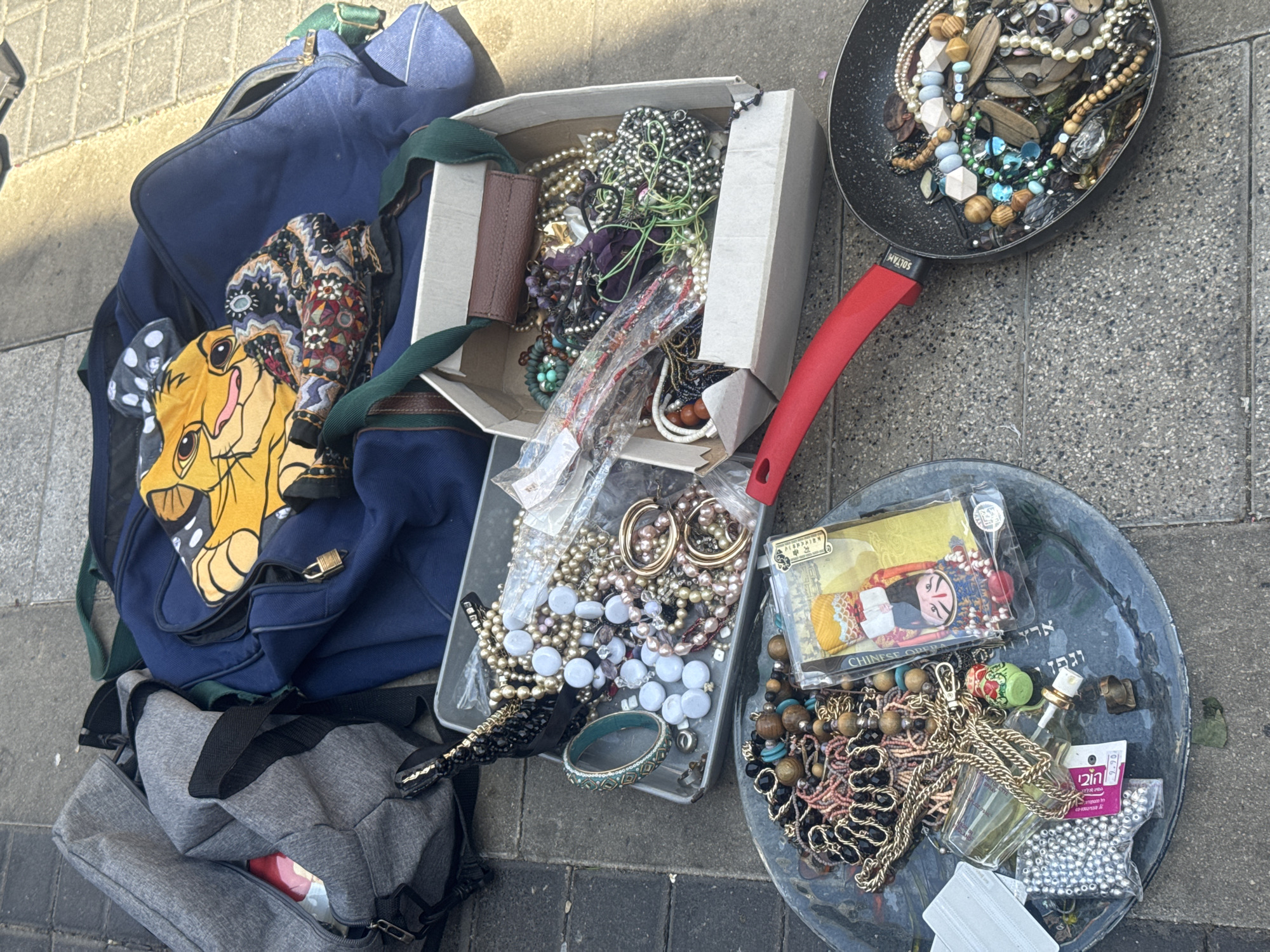 The image shows a collection of items spread out on a paved surface, likely a sidewalk. There are two bags, one blue with a "Lion King" character patch, and another gray. Additionally, there is a cardboard box and two pans, one black with a red handle and another gray. Each container holds a variety of jewelry items. The scene appears to be set in an urban environment, potentially a flea market or street sale. The bags and boxes are open, suggesting they are being displayed for sale. The presence of Hebrew writing on a label in the gray pan indicates the location is likely in Israel, possibly in Tel Aviv, based on similar scenes commonly found in the city's markets and street sales. There are no people visible in the image. The focus is on the objects and their arrangement on the ground.