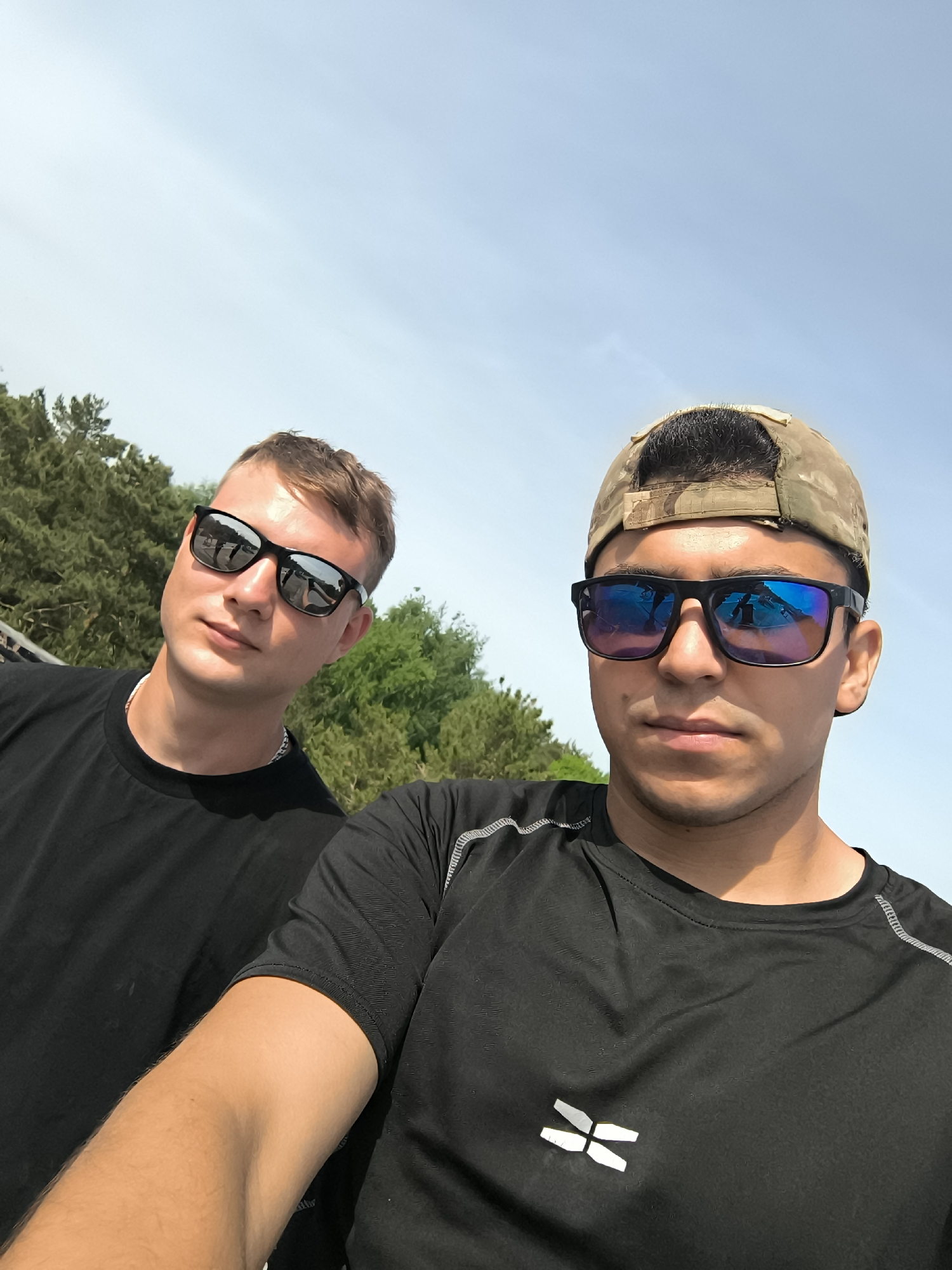 Two young men pose for a selfie against a backdrop of trees and a bright blue sky. The man on the right wears a camouflage baseball cap, a black athletic shirt with a logo, and blue mirrored sunglasses. The man on the left is wearing a black t-shirt and black sunglasses. Both men appear to be outdoors in a sunny, natural environment. The city and specific location are undetermined.