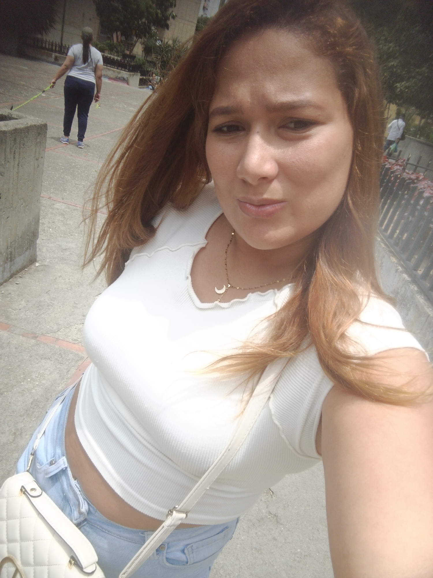 A woman with fair skin and auburn hair, wearing a white ribbed crop top and light blue jeans, takes a selfie. She is wearing a gold necklace with a crescent moon pendant, and a white quilted purse is slung over her shoulder. In the background, another woman wearing a grey shirt and dark pants walks a dog on a leash. A man in a white shirt can also be seen in the distance. The location appears to be an outdoor urban area, possibly in the city of Caracas, Venezuela, as per the prompt.