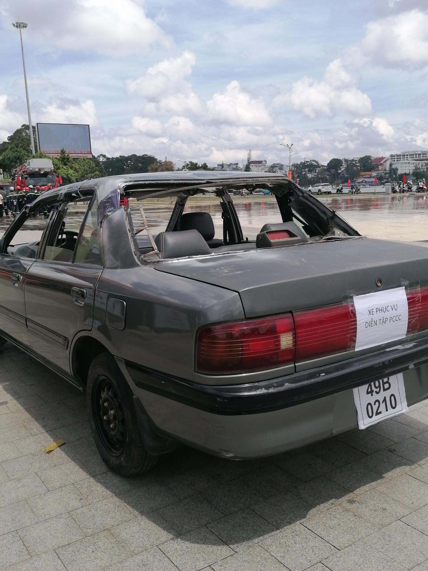 The image shows a severely damaged grey sedan, likely a Mazda, with its roof and windows completely shattered, positioned on a paved outdoor area. The car's interior, including seats, is visible through the open top. A white piece of paper with Vietnamese text, "XE PHỤC VỤ DIỄN TẬP PCCC," is taped to the rear of the car, indicating it's for fire drill purposes. Below it, a license plate reads "49B 0210." In the background, a large screen is visible, along with a tall light pole and several fire trucks with emergency personnel in uniform. The sky is cloudy. The scene is set in Da Lat, Vietnam, as indicated by the license plate format (49 is the code for Da Lat province). The ground appears wet, suggesting it may have recently rained.