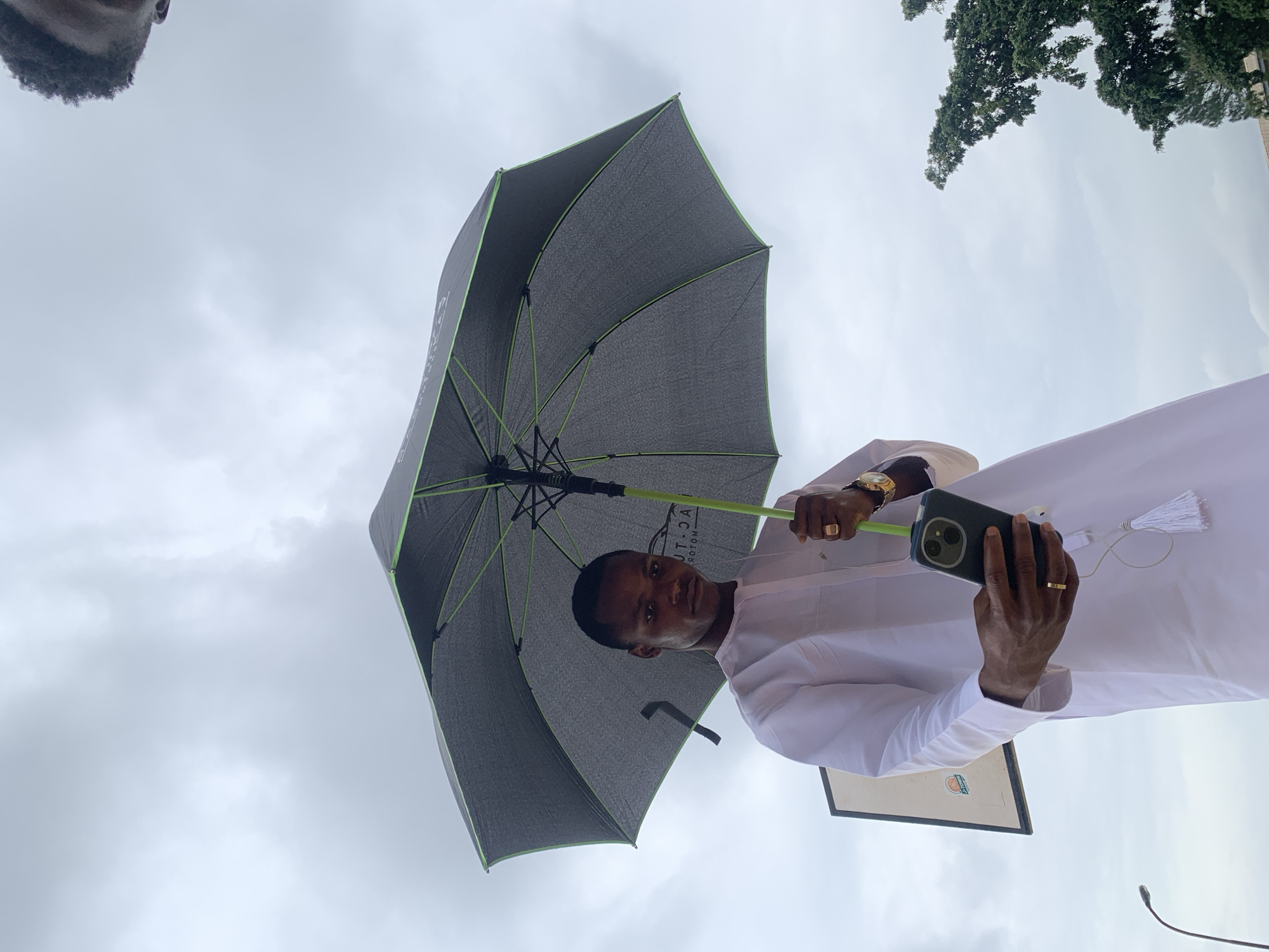 A low-angle, full-body shot captures a young man standing outdoors under an open, dark gray umbrella with a neon green trim and handle. He is dressed in a white traditional outfit, possibly a boubou or agbada, with a tassel detail hanging from his right side. He is looking at his smartphone, which he holds in his right hand. On his right wrist, he wears a gold watch, and a gold ring is visible on his right ring finger. The background consists of an overcast sky and the top of a green tree on the right side of the frame. A sign with text and a graphic of a car is partially visible to the left of the man. The scene suggests an outdoor setting in Jos, Nigeria, likely during a cloudy or possibly rainy day, given the open umbrella.