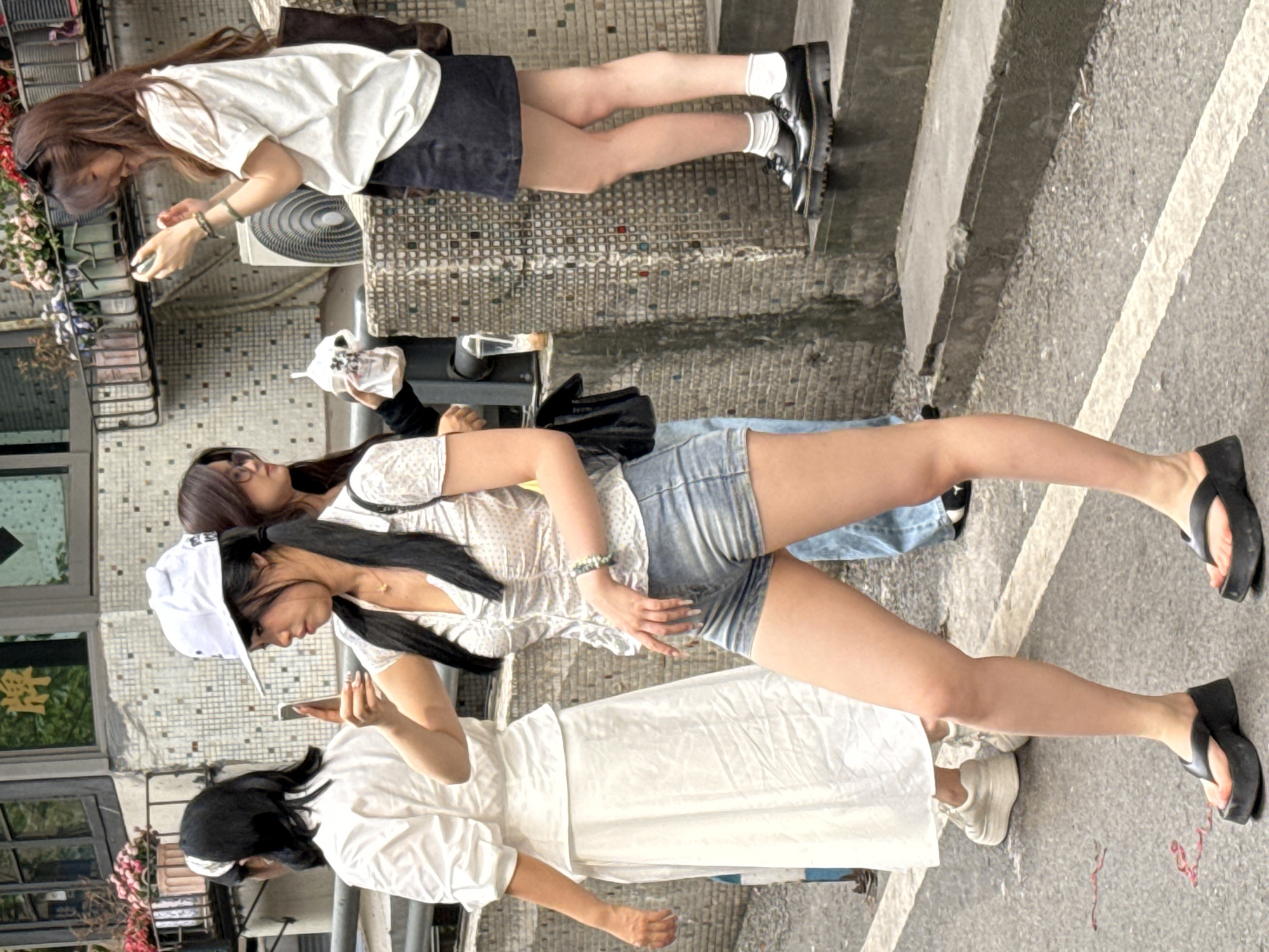 The image shows three young women standing on a paved surface in an urban setting. The backdrop includes buildings with windows and balconies. A young woman wears a white t-shirt, a navy-blue skirt, white socks, and black boots. A second young woman wears a white patterned top and denim shorts. The third young woman wears a white dress. They are all looking at their phones. Based on the buildings, the location is most likely Yuzhong District, China.