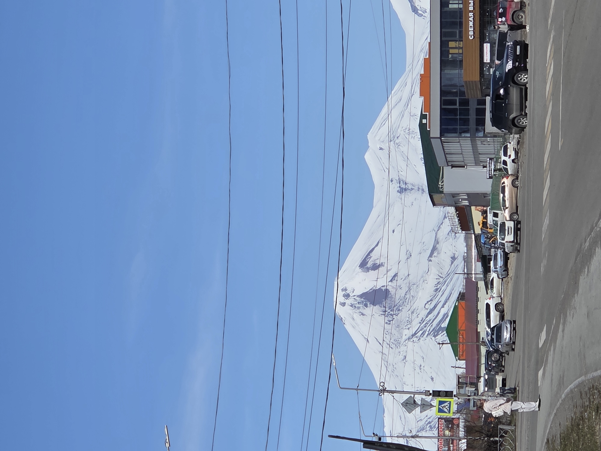 The image captures a street scene in an unidentified city. A snow-capped mountain dominates the backdrop, partially obscured by overhead power lines. Along the right side of the frame, a row of parked cars lines the edge of a paved road. Buildings, some with visible signage in a non-English script, stand adjacent to the parking area. A pedestrian wearing light-colored clothing walks along the roadside. A pedestrian crossing sign is visible. The sky above is clear blue.