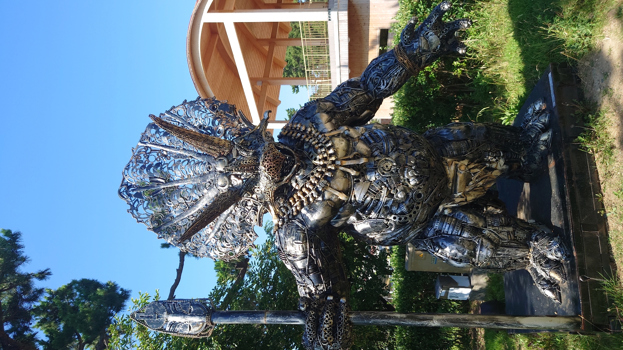 This is a full-shot, eye-level, outdoor, daytime photograph of a large, metallic sculpture resembling a dinosaur or a creature with dinosaur-like features. The sculpture is made of various recycled metal parts and is situated in a grassy area with some bushes in the background. To the right of the sculpture, a modern, open-air building with a wooden roof structure and a railing is visible. The sky is clear and blue. The sculpture is dark, silver, and metallic in color, with intricate detailing, including a large frill on its head, sharp horns, and a beaded necklace. It is standing on a dark platform. The sculpture is holding a large spear in its left hand. The image is located at the Ulsan Grand Park, in the city of Ulsan, South Korea.
