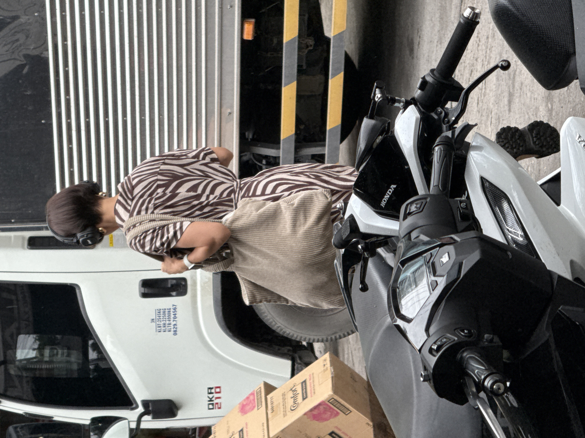 The image depicts an outdoor scene, likely on a street in Nha Trang, Vietnam, as suggested by the presence of motorcycles and a truck common in urban Vietnamese environments. In the foreground, two motorcycles are parked, with one partially visible in the lower right corner. The primary focus is on a woman standing beside a white truck. She is wearing a zebra-print dress, headphones, and carrying a large, light-brown tote bag. The truck has several markings on its side, including "QKR 210" and a series of numbers and letters that appear to be vehicle identification or load information. Behind the woman, the corrugated metal side of the truck's cargo container is visible. Cardboard boxes, one labeled "Comfort," are stacked to the left of the truck. The ground is paved, suggesting a public thoroughfare or parking area. The overall impression is of a mundane urban moment, potentially involving the loading or unloading of goods.