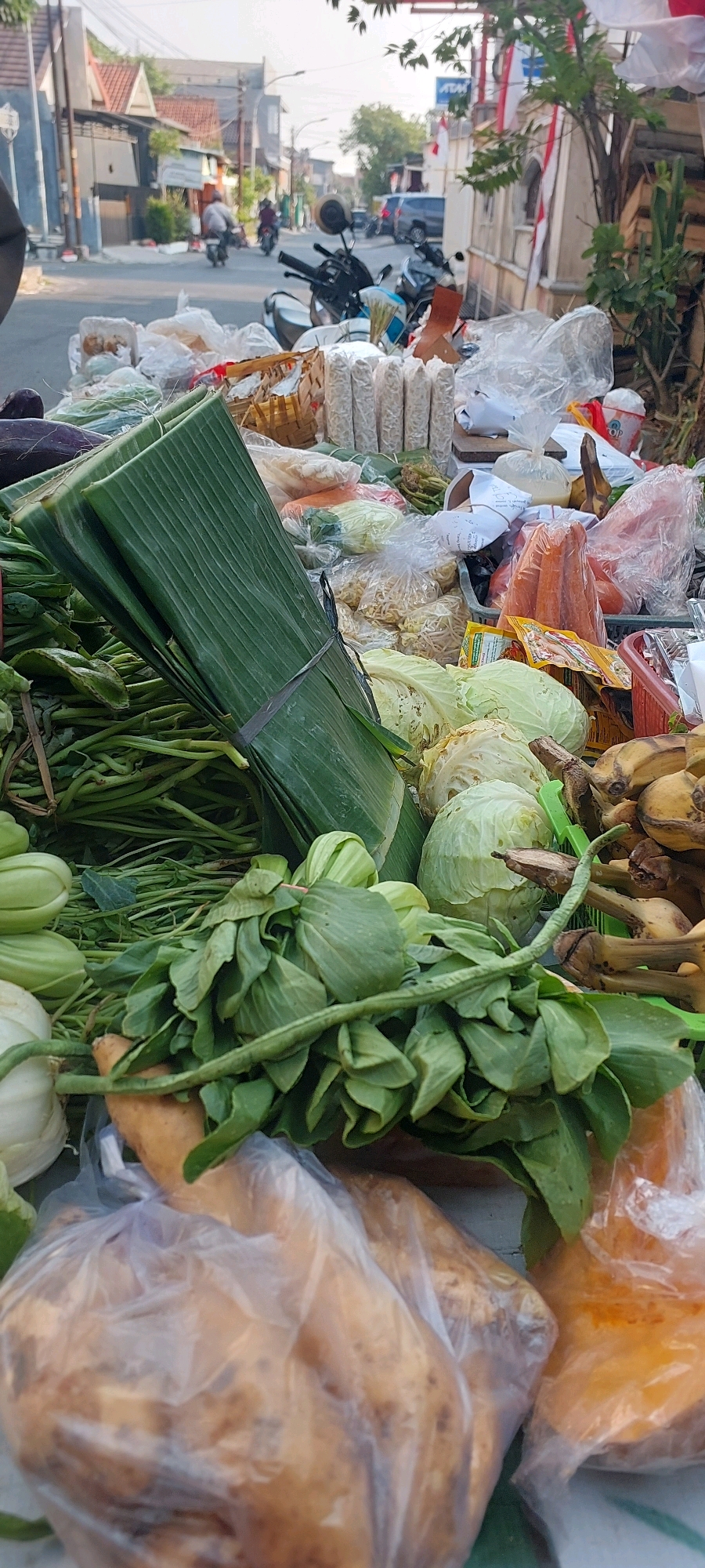The image depicts a street-level view of a produce stall in Semarang, Indonesia. A variety of fresh vegetables are displayed, including bunches of green leafy vegetables, cabbages, bananas, and what appear to be sweet potatoes packaged in plastic. In the background, a street is visible with several motorcycles and cars. Buildings line the street, and a few individuals can be seen in the distance. The overall scene suggests a typical market environment within the city.