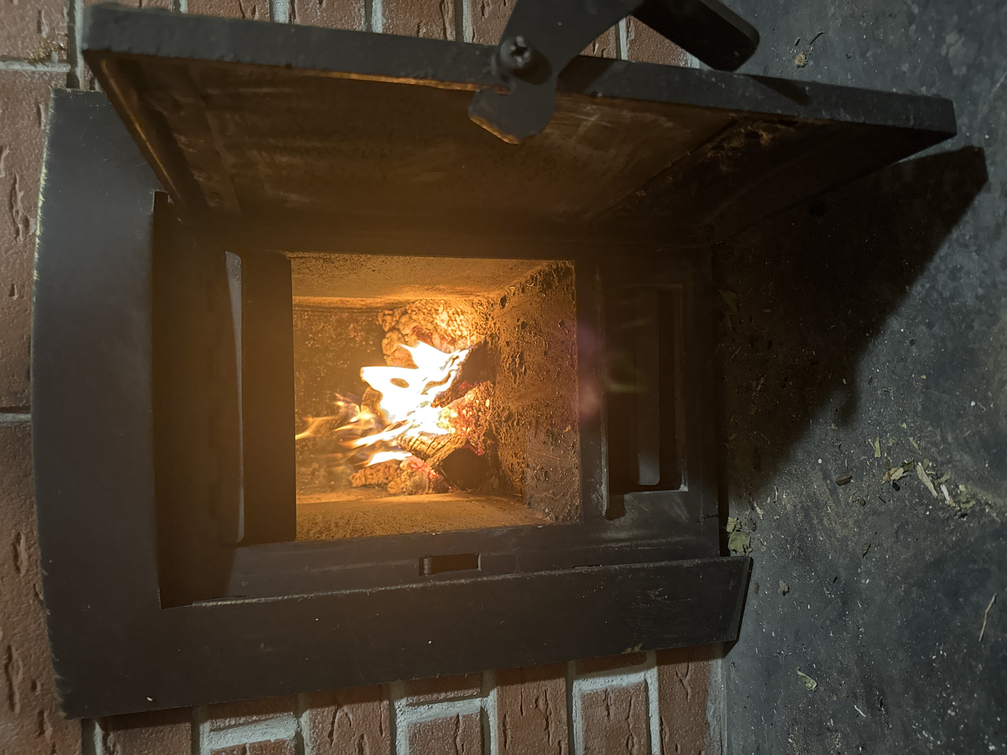 A close-up shot shows a brick fireplace with a black metal door partially open, revealing a fire burning inside. The door has a handle at the top. The fireplace is set into a brick wall. Debris is scattered on the floor near the fireplace. The location is not discernible from the image.