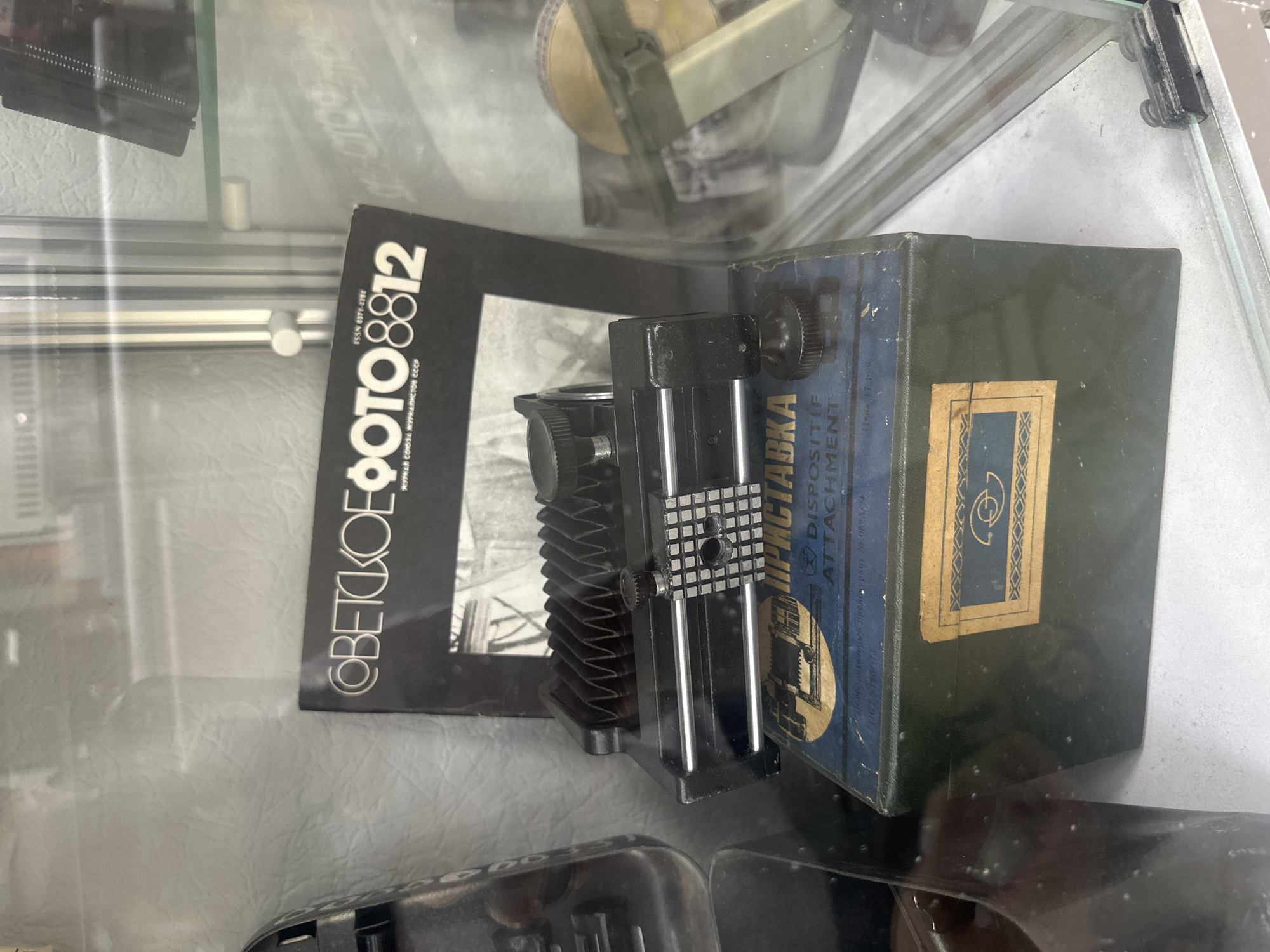 The image shows a display case with vintage photographic equipment. In the foreground, there is a bellows attachment for a camera, resting on a green box with Cyrillic text. Above the bellows is a magazine titled "Советское Фото" (Soviet Photo) with the issue number 8812. The display case appears to be in a museum or a shop. The overall scene is well-lit, suggesting an indoor environment. No people are visible in the image. The specific location in Tobolsk, Russia, is not discernible from the image content.
