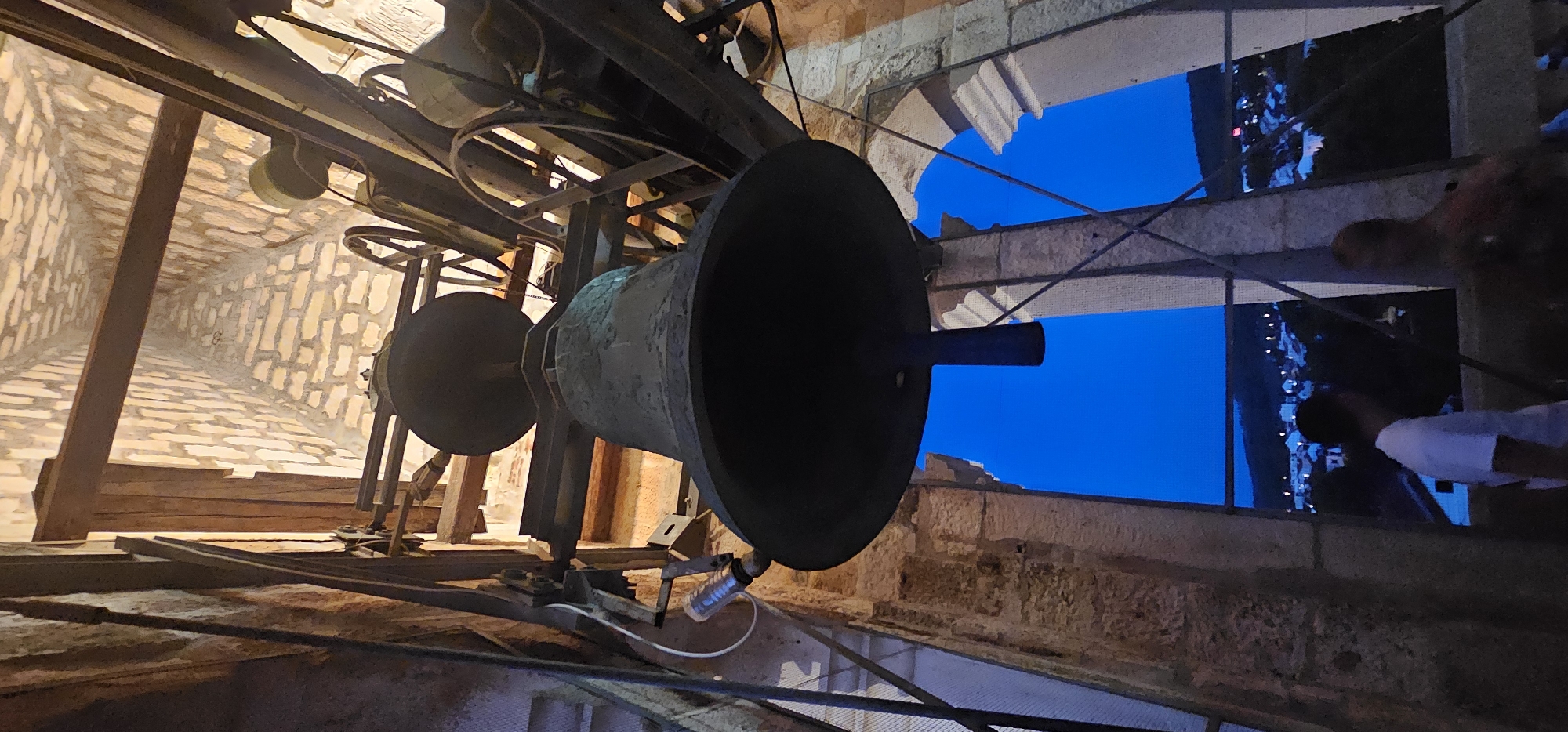 An extreme low-angle, vertical medium shot captures the interior of a bell tower in Supetar, Croatia, at dusk. Dominating the frame is a large, dark bell, silhouetted against the deep blue twilight sky visible through arched openings. The bell is suspended from a wooden and metal structure with visible pulleys and mechanical components, suggesting its operational status. To the left of the bell, a white pipe with a coiled cord is attached to the structure.
In the foreground, two individuals are partially visible. One, wearing a white t-shirt, stands on the left, facing away from the camera and towards the openings. Another figure, out of focus and further to the right, is also partially visible. Through the openings, a distant view of a town with illuminated lights is discernible, nestled against a dark hillside. The architecture of the tower is characterized by rough-hewn stone walls and wooden beams, with a high, vaulted ceiling constructed of light-colored bricks.