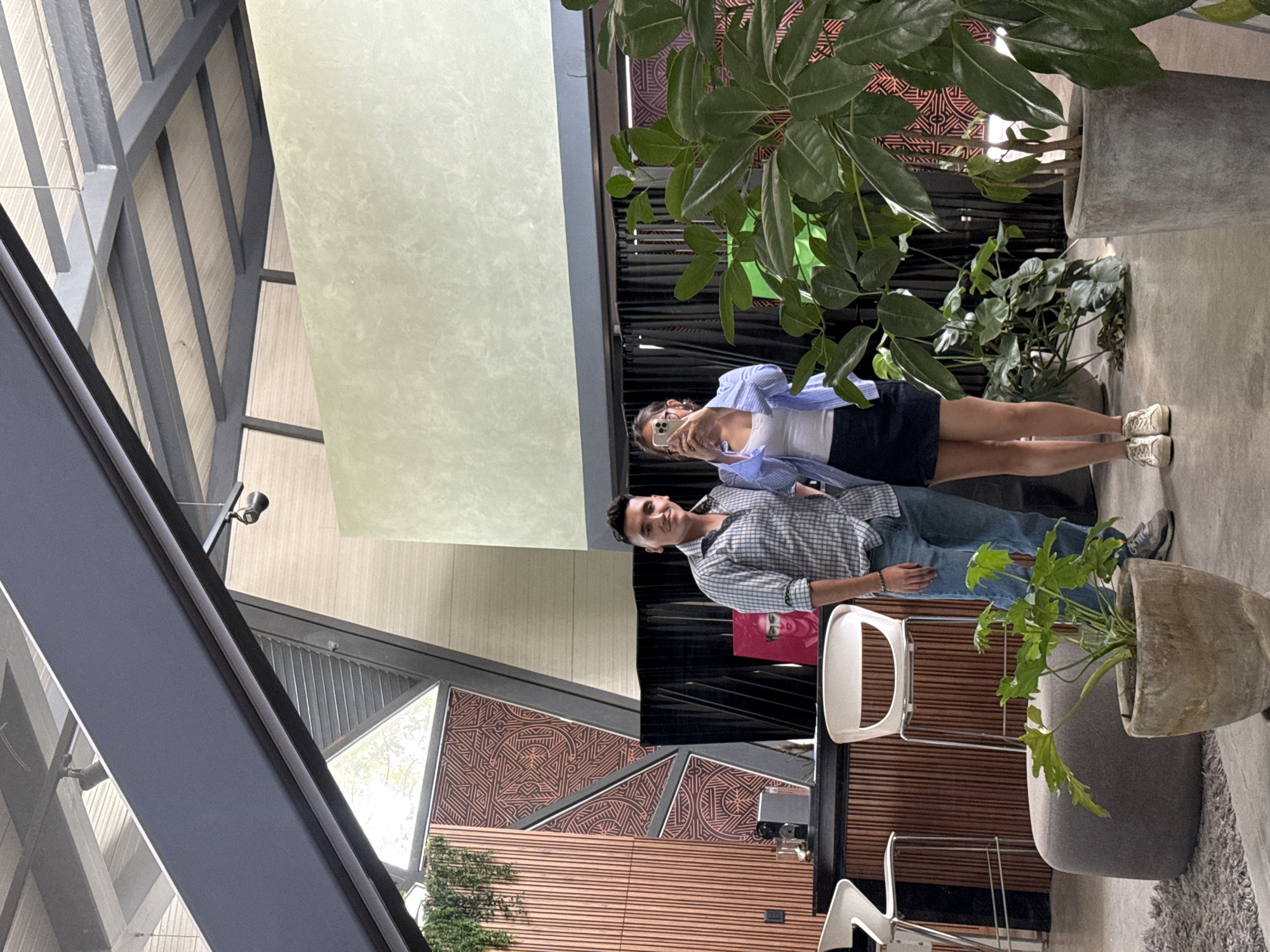 The image depicts a man and a woman standing in a modern indoor space. The man is wearing a light blue and gray checkered shirt, blue jeans, and gray sneakers. He is looking toward the camera. The woman is wearing a light blue shirt, a white tank top, and a navy blue skirt. She is holding her phone up, seemingly taking a picture.
Behind them is a wall with brown and gray geometric patterns. To the right of the couple, there are large potted plants. There is a white bar stool near the man. The space appears to be a cafe or a modern building with a view of a landscaped area outside.
The location is San Bernardino Tlaxcalancingo, Mexico.