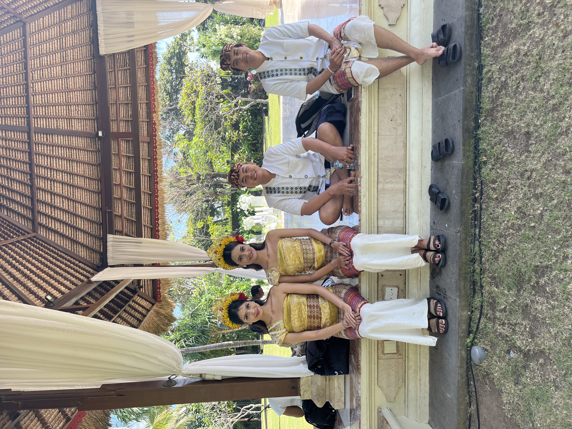 Four individuals are seated on a raised stone platform beneath a thatched-roof pavilion with white drapes. Two women, positioned on the left, wear traditional Balinese attire consisting of ornate gold and yellow tops, patterned waist sashes, and white lower garments. They also wear elaborate headpieces adorned with yellow flowers and red accents. Two men, seated to the right, are dressed in white shirts with patterned collars and bands, and traditional Balinese headbands. One man is wearing a white lower garment, while the other is wearing a patterned sash over a white lower garment.
In front of the seated individuals on the stone platform, there are several pairs of black flip-flops and a plastic water bottle. Bags are also visible near the seated individuals. The background reveals lush green foliage, including palm trees, and a bright blue sky, suggesting an outdoor tropical setting. The architecture of the pavilion, with its thatched roof and wooden beams, is characteristic of South Kuta, Indonesia, where such structures are common for outdoor events and hospitality venues. The overall scene appears to be a staged photo opportunity, possibly related to a cultural event or wedding.