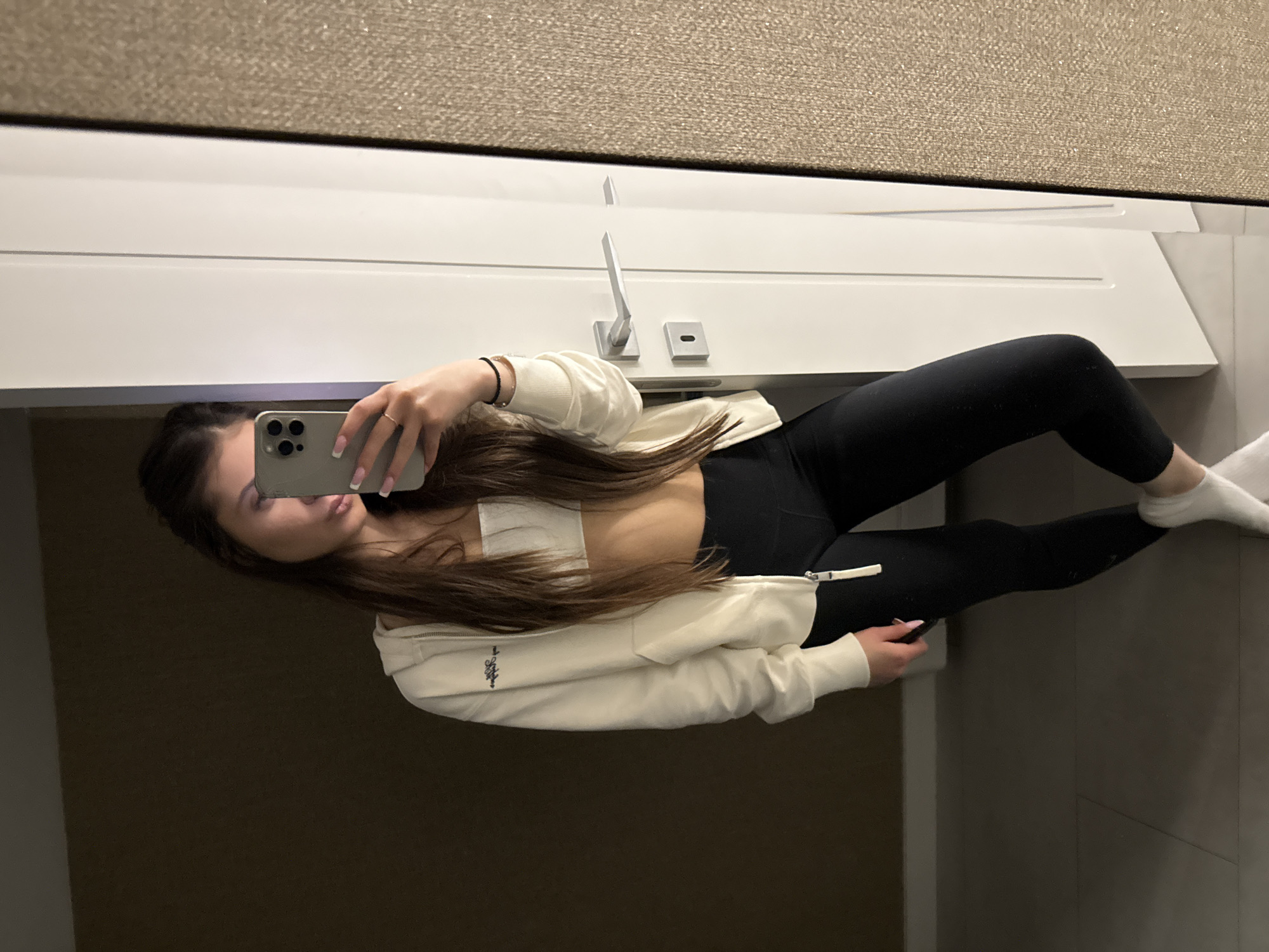 A young woman is taking a selfie in a bathroom. She's wearing a white sports bra, black leggings, a white zip-up hoodie, and white socks. She is holding a smartphone in her left hand. The background shows a white sink counter with a modern faucet, and a wall covered in a beige textured material. The location cannot be determined from the image.