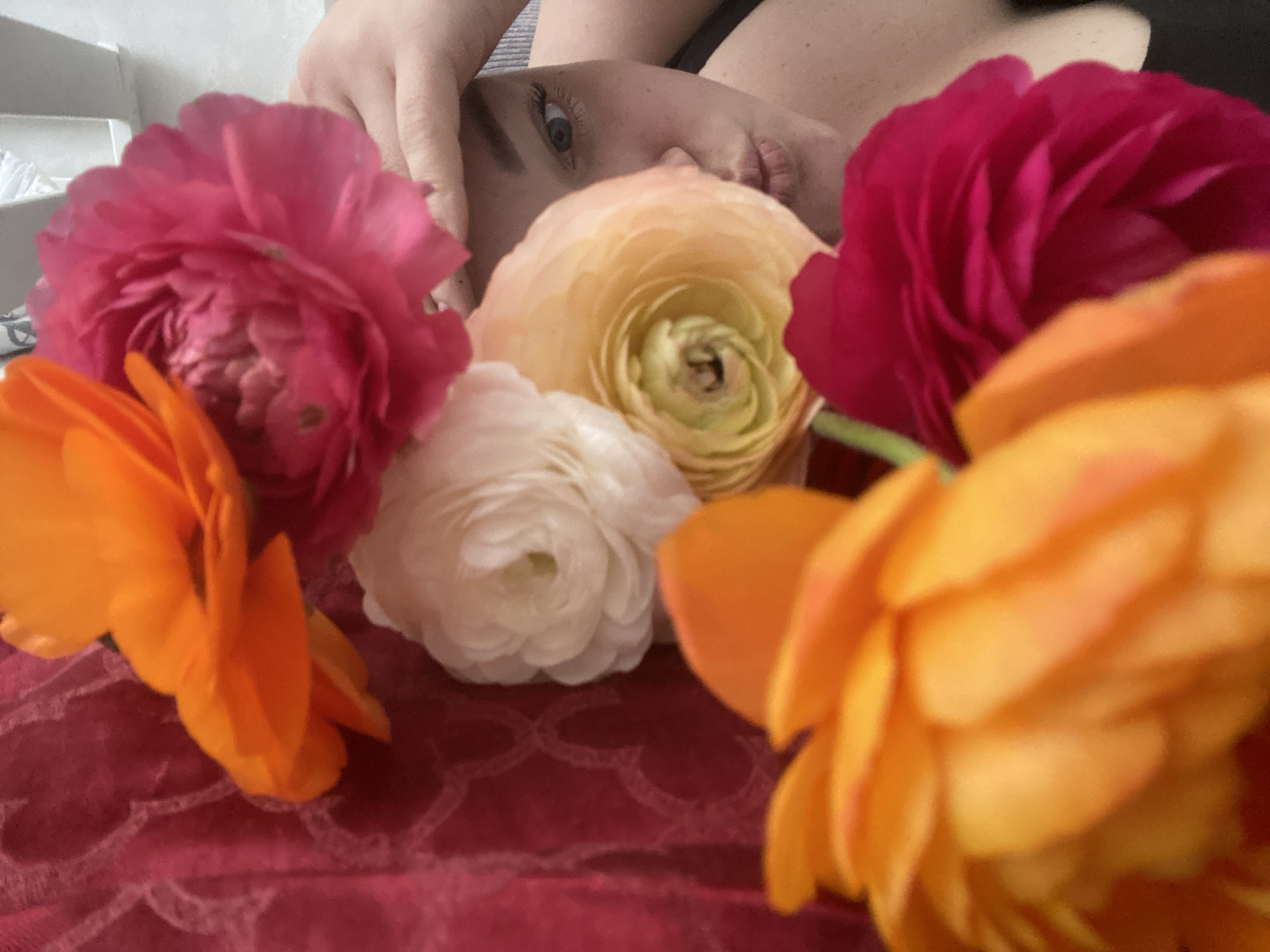 This close-up image, reportedly located in Sochi, Russia, features a person lying down, their face partially visible and framed by colorful flowers. The person's left eye, part of their nose, and lips are discernible in the upper-middle section, with a hand resting near their cheek. The visible eye has a blue or green iris, and the expression appears serene or contemplative.
The foreground and midground are filled with an abundance of ranunculus flowers in various vivid colors, including deep magenta, light yellow, white, and bright orange. These flowers are arranged around the person's face, creating a soft, natural frame. Some flowers are in sharper focus, particularly the yellow one in the center and the white one below it, showcasing their intricate, ruffled petals. The orange flowers are prominent in the lower left and right foreground, appearing slightly blurred.
The person and flowers rest on a dark pink or red fabric with a subtle, lighter-toned patterned overlay, possibly a lace or embossed design, visible in the lower half of the image. The background is softly blurred, showing lighter elements on the left, possibly furniture or a wall, and darker tones on the right. The overall setting appears to be indoors, likely a bedroom or a cozy lounge area.
The image conveys a gentle and aesthetically pleasing mood, emphasizing natural beauty and tranquility. There is no visible text, and the specific time of day or weather conditions are not discernible from the indoor scene.