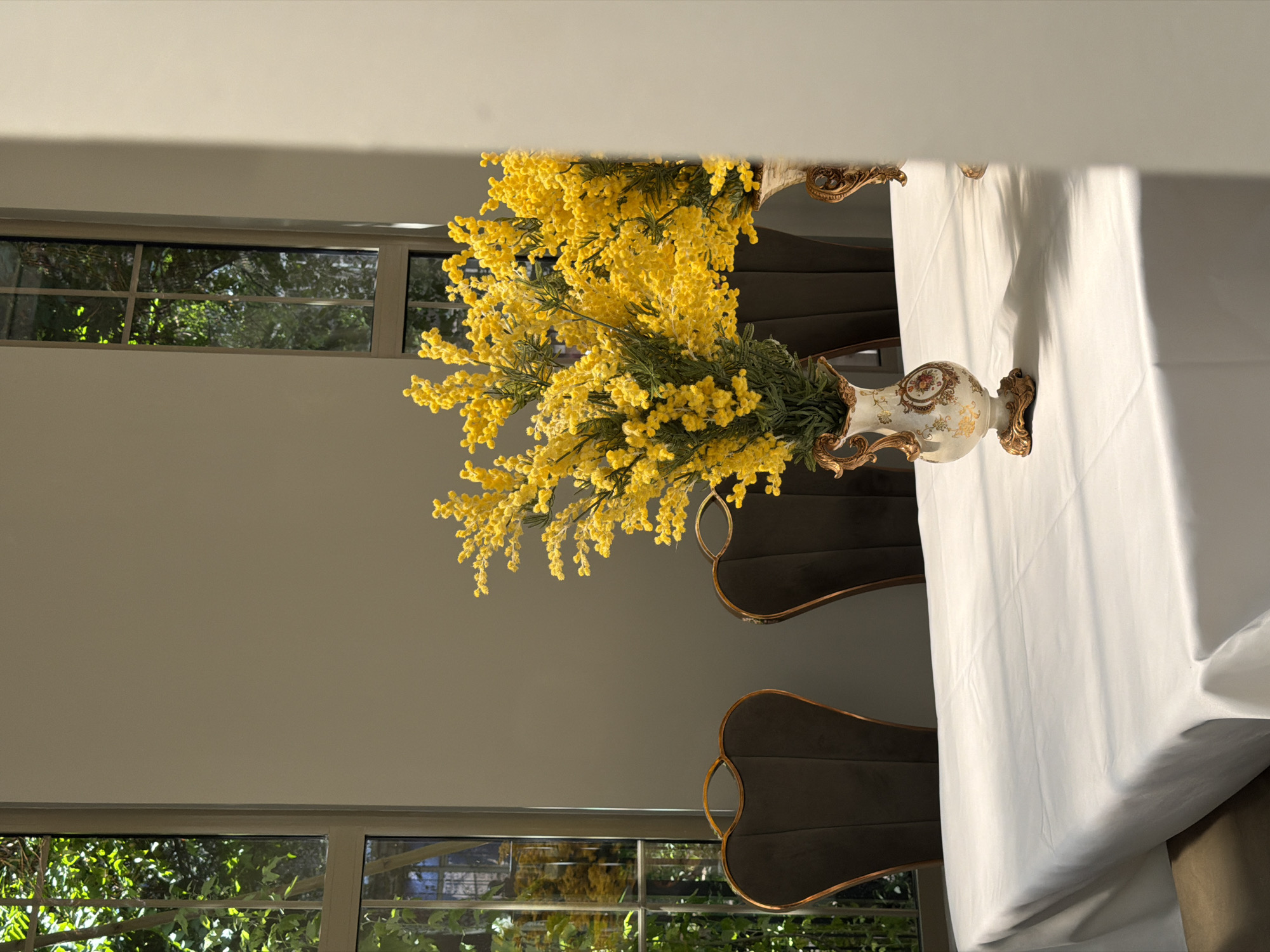 A vase of yellow flowers sits on a table covered with a white tablecloth, inside a building. There are chairs surrounding the table. Outside of a window, greenery and trees are visible. There are no people visible in the scene. Location is indiscernible; there are no specific elements identifiable as being in Bishkek City, Kyrgyzstan.