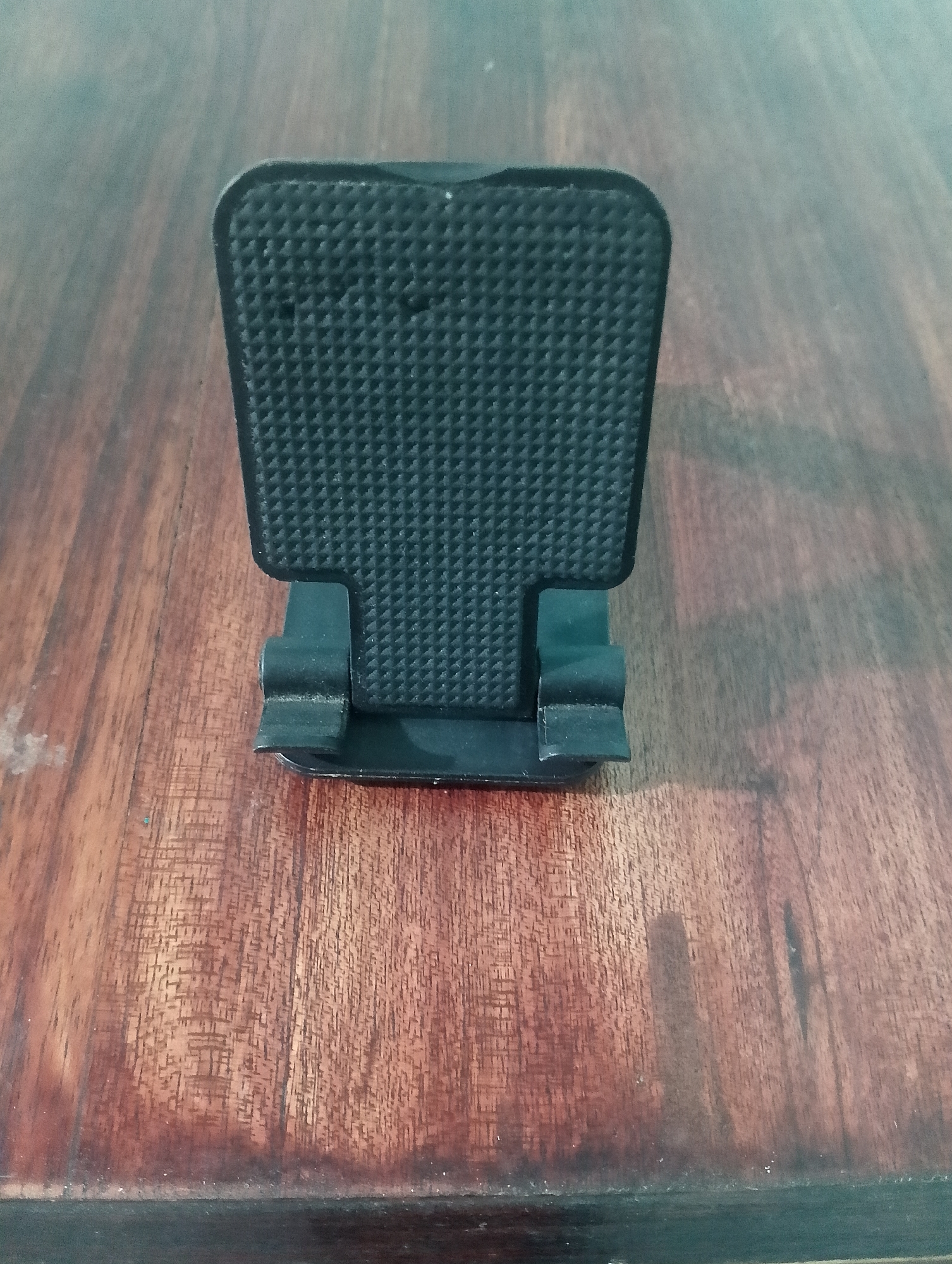 A black, textured foot pedal rests on a dark wooden surface. The pedal is rectangular with rounded corners and a diamond-shaped pattern on its surface. It is attached to a black base with two angled supports. The wooden surface has a visible grain pattern and appears to be a table or desk. The background is blurred, suggesting a shallow depth of field. There are no people or other objects clearly visible in the image. The scene is indoors and the lighting is diffuse. The specific location in Serbelawan, Indonesia is not discernible from the image.