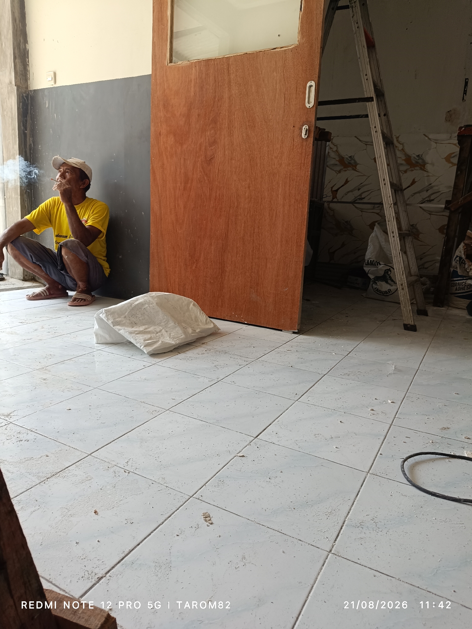 A man wearing a yellow t-shirt and dark shorts is crouched on a tiled floor, smoking a cigarette. He is leaning against a gray wall with a wooden door to his right. In the background, a ladder is propped against the wall, and several bags of construction material are visible. The floor is dusty, and there are some debris scattered around. The scene appears to be an indoor construction or renovation site, possibly in Sruni, Indonesia. A date and time stamp at the bottom right corner indicates "21/08/2026 11:42".