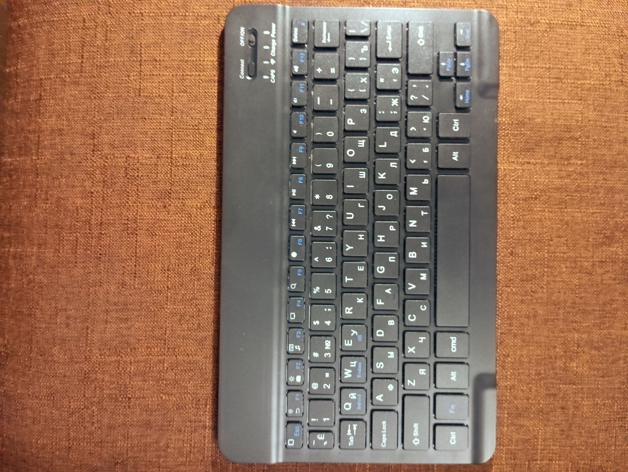 This is a top-down, eye-level shot of a black wireless keyboard resting on a textured, reddish-brown fabric surface. The keyboard is oriented vertically in the frame, with its top edge closer to the viewer. It appears to be a compact, full-sized keyboard with standard QWERTY layout on the left side and Cyrillic characters on the right.
Prominent keys include:
- On the left side, a vertical row of modifier keys: Ctrl, Alt, Fn, Shift, and Caps Lock.
- The standard alphanumeric keys are visible, with English letters on top and Cyrillic letters below.
- A number row is present above the letter keys.
- Function keys (F1 through F12) are located along the right edge of the keyboard.
- Special keys like Esc, Tab, Enter, Spacebar, and navigation keys (arrow keys, Home, End, Page Up, Page Down) are also visible.
- Along the bottom right edge, there are three small switches labeled "Connect," "Off/On," and "Caps."
The fabric surface has a coarse weave and exhibits a slight sheen, suggesting it could be upholstery or a similar textile. The lighting is even, casting minimal shadows, which allows for clear visibility of the keyboard's details. The image is sharp and in focus throughout the keyboard area.
There are no people or animals present in the image. The scene is sterile and lacks any context of location or activity beyond the object itself. The image is a straightforward product shot of a keyboard.