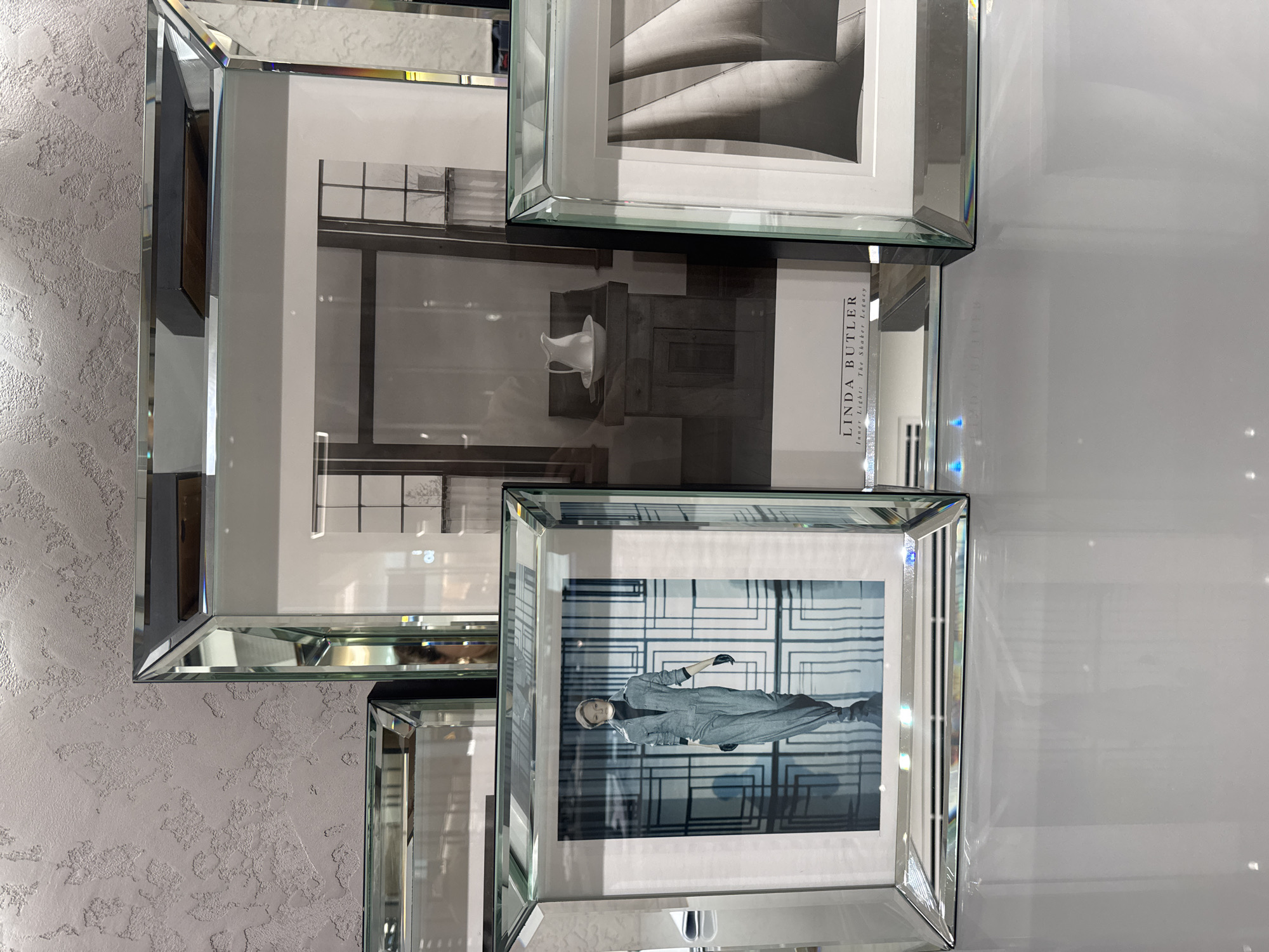 The image shows a decorative arrangement of framed black and white photographs on a wall. The frames appear to be mirrored. The visible photographs depict a staircase, a still life with a white object (possibly a pitcher) and what looks like the corner of a room, and a person in a jumpsuit posed in front of a grid-like background. A logo "LINDA BUTLER" along with "Inner Light: The Shaker Legacy" is visible on one frame. The left wall has a textured, grey finish while the right wall is smooth and white. The image, due to the logo, could be from a place offering Linda Butler's photography, possibly within the city of Dubai, United Arab Emirates.