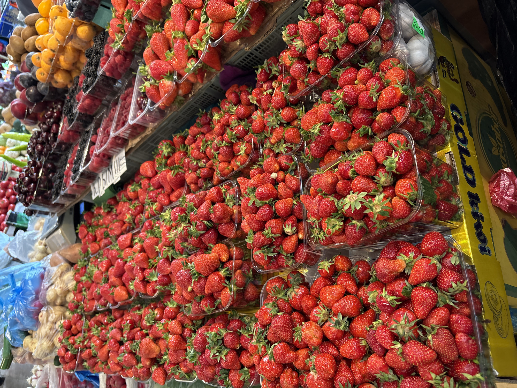 A display of fruits, with a dominant presence of strawberries in clear plastic containers, likely at a market or stall in город Ялта, Ukraine. Other fruits like cherries, plums, raspberries, and blackberries are also visible, but are less prominent. To the left are garlic and other produce.