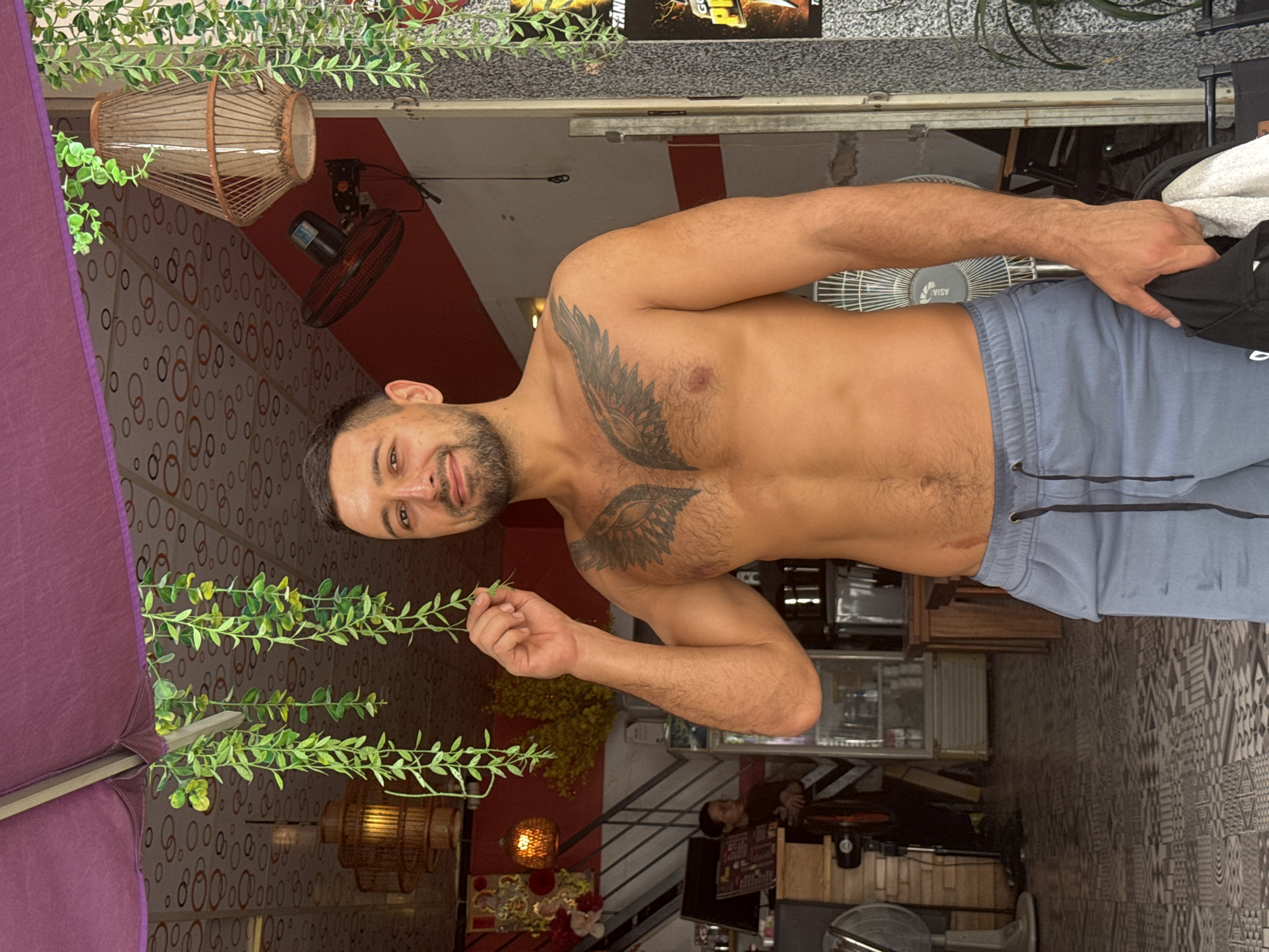 A shirtless man with a winged tattoo on his chest stands in front of an open-air establishment. He is wearing light blue athletic shorts and holding a black bag with a white towel. In the background, an interior space with shelves, coolers, and a person can be seen. Hanging plants and a decorative lantern are visible overhead. The man is holding a green leaf up to his face. The scene appears to be in a warm, tropical climate, possibly a cafe or shop in Nha Trang, Vietnam, given the architecture and foliage.