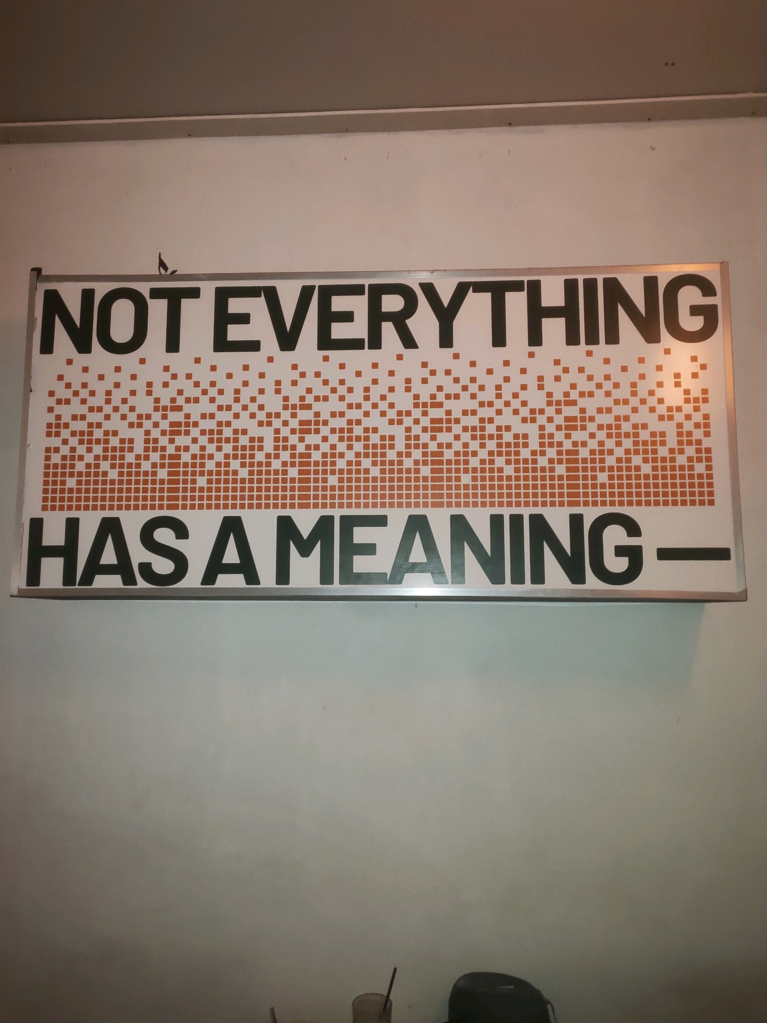 A rectangular sign is mounted on a light-colored wall. The sign has a silver frame. The top line of text on the sign reads "NOT EVERYTHING" in large, dark sans-serif font. Below this text is a pattern of small orange squares forming a gradient from solid to sparse, from left to right. The bottom line of text on the sign reads "HAS A MEANING-" in the same large, dark sans-serif font as the top line, with a horizontal line extending to the right after the word "MEANING." The wall itself is plain and appears to be indoors, with a darker ceiling visible at the top of the frame. In the lower portion of the image, the wall transitions to a lighter shade. A few objects are visible at the bottom edge of the frame, including what appears to be a glass with a straw and a dark, rounded object. The context of the city of Pagerwojo, Indonesia, is not evident from the visual information in the image.