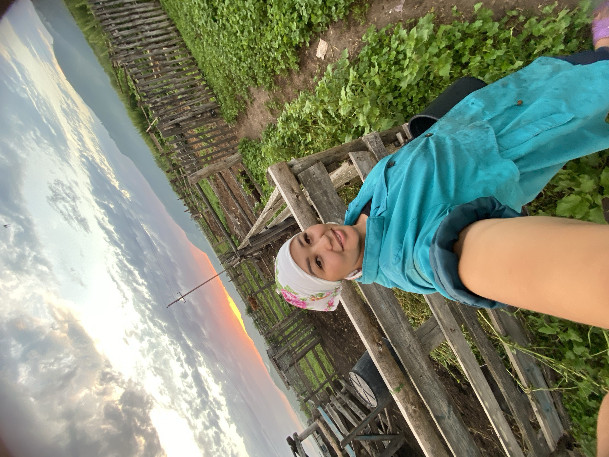In an outdoor scene located near Maloarslangulovo, Russia, a woman is captured in a selfie. She is wearing a white headscarf with a floral pattern and a turquoise jacket. She appears to be standing in a fenced enclosure, possibly a corral or farmyard, as indicated by the wooden fence and rough ground visible. To the left, part of a wooden structure and a large black barrel are present. The background features a rolling landscape with lush green vegetation and distant mountains. The sky is dramatic with a mix of clouds and a striking sunset casting a warm glow on the horizon. A single cow can be seen in the distance within the enclosure. The overall impression is one of rural life during dusk.