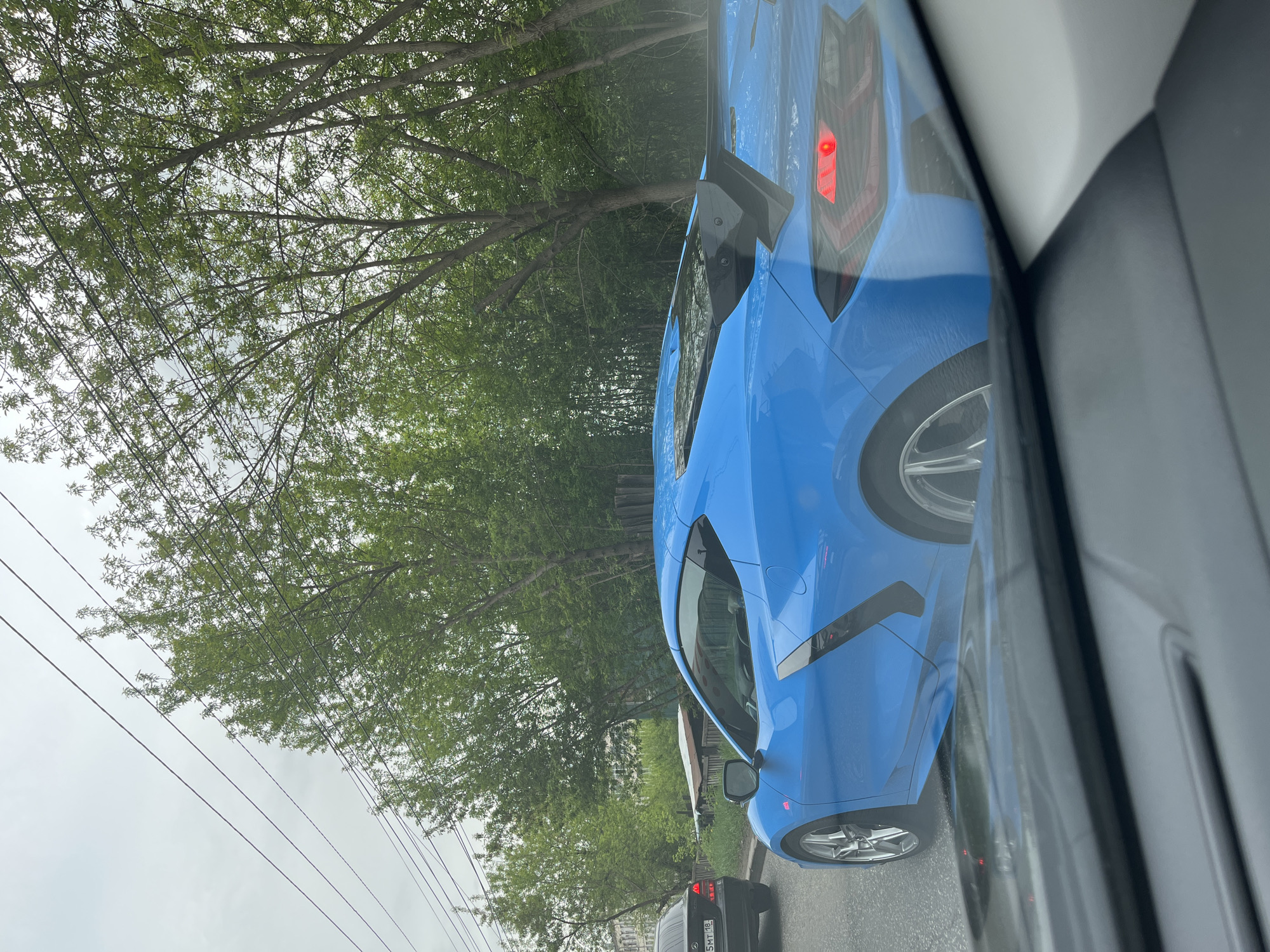 The image depicts a blue sports car with its brake lights illuminated. It appears to be a Corvette. The car is positioned on a road, likely in traffic. There is a black car in the lane behind it. Trees and overcast sky are visible in the background. The photo was taken from within another vehicle. The car's license plate is partially visible but not readable. According to the license plate of the car following, this is likely the city of Izhevsk, Russia.