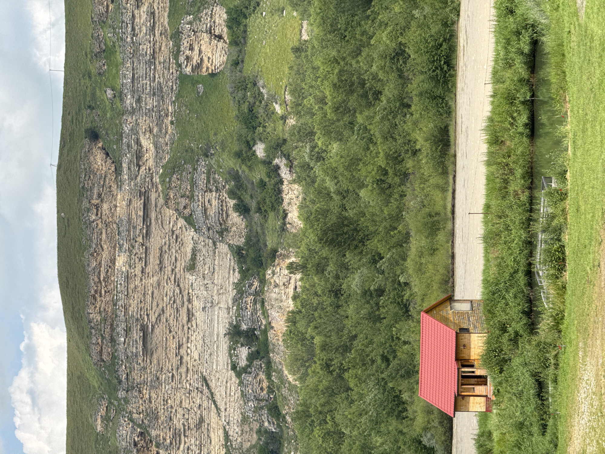 A small, rustic wooden house with a red tiled roof sits on a grassy bank beside a muddy river. The house has a simple, rectangular design with a porch. Behind the house, a dense forest stretches across the landscape, and further in the background, a massive, layered cliff face rises dramatically. The cliff is composed of light-colored rock with distinct horizontal striations, and it is partially covered in green vegetation. The sky above is partly cloudy with scattered white clouds against a pale blue backdrop. The scene depicts a remote, natural setting.