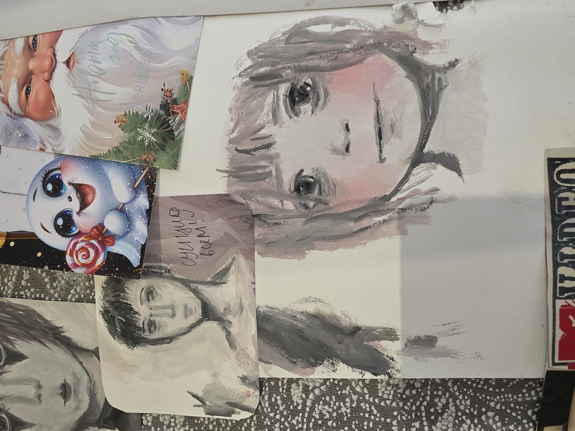 The image shows an overhead view of a surface covered with various items. At the top left is a Christmas card featuring Santa Claus. Adjacent to it is another Christmas card with a snowman holding a lollipop. Below these are several grayscale portrait drawings of people. The portraits appear to be painted on paper. The background surface has a patterned texture. A portion of a sticker with the word "VIDEO" is visible on the right side. The location of Chaikovsky, Russia, cannot be determined from the image alone.