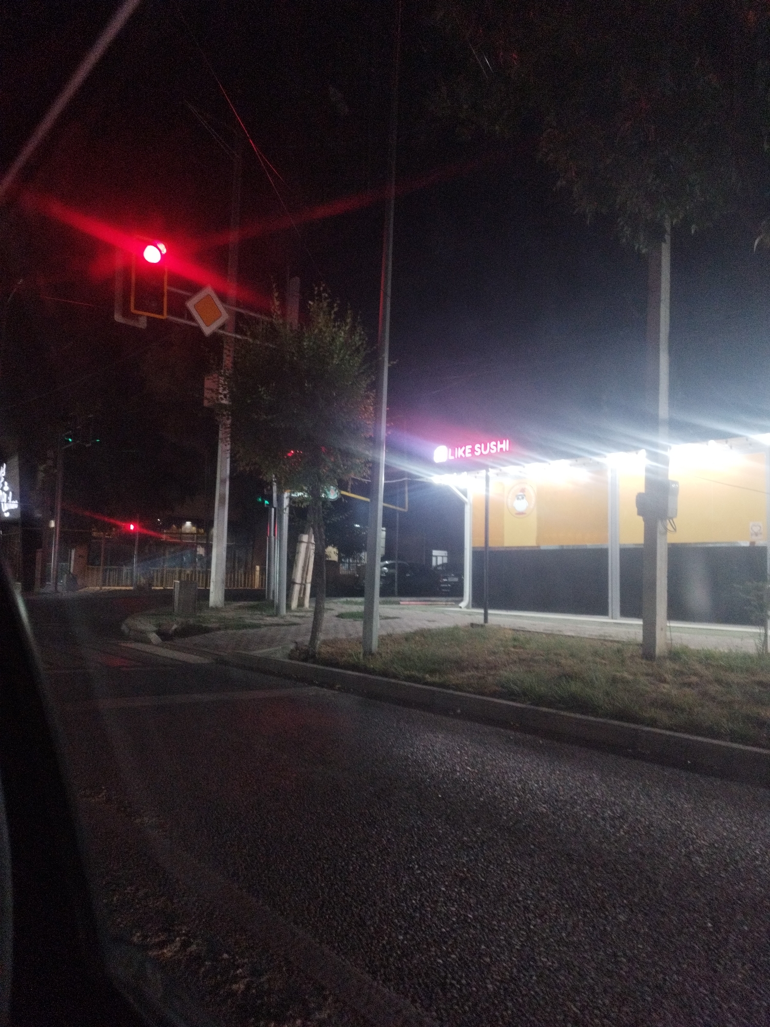 An image taken at night on a city street in Shymkent, Kazakhstan shows a traffic light displaying a red signal, a diamond-shaped yield sign, and a neon sign that reads "LIKE SUSHI" above a building with yellow and black panels. The street is paved and appears wet, with some grass and trees lining the sidewalk. Power lines are visible overhead. The perspective suggests the photo was taken from inside a vehicle.