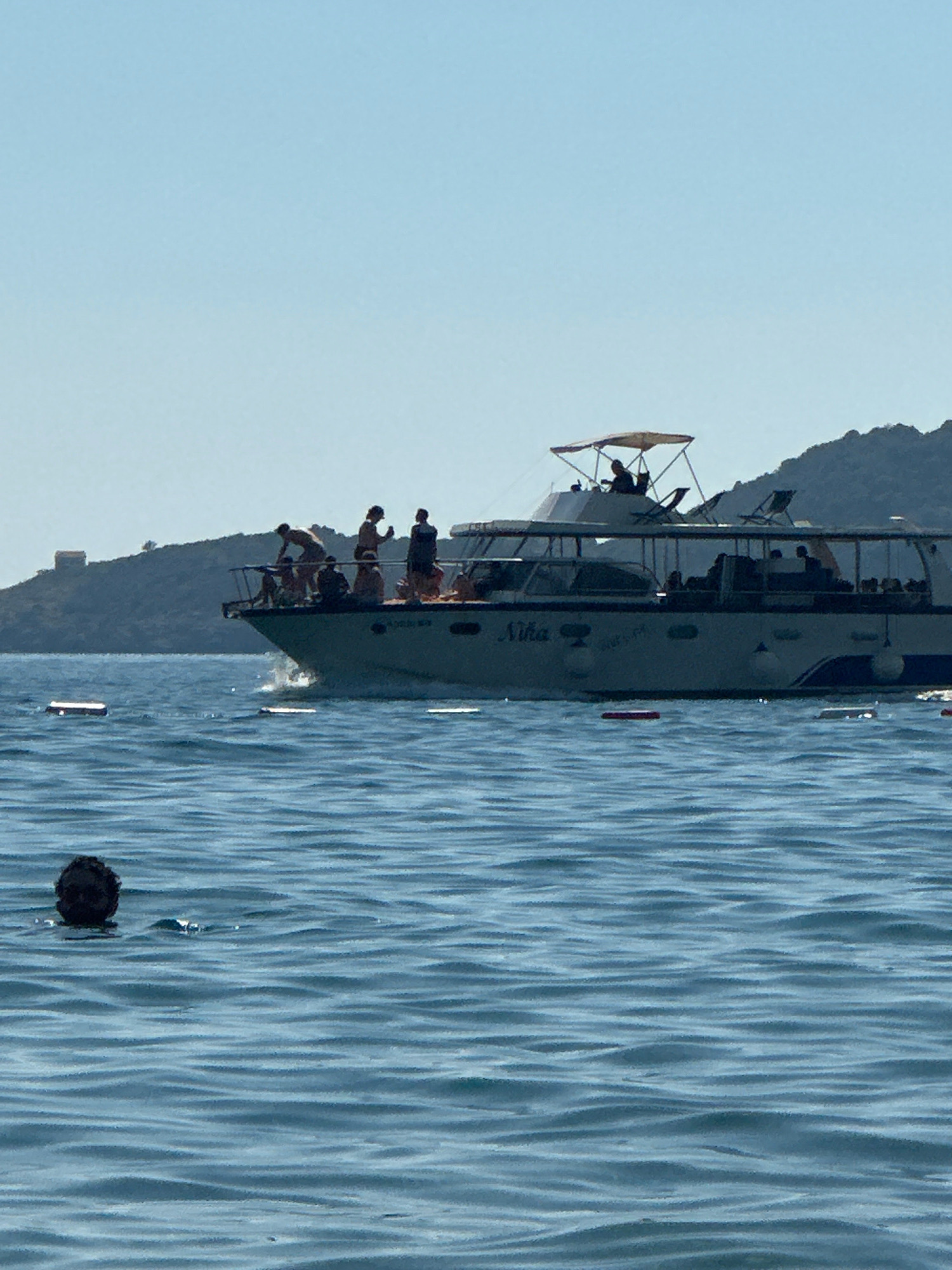 The image depicts a sun-drenched day with a clear blue sky over the water. In the foreground, a person's head is visible above the water, appearing to be swimming. The middle ground is dominated by a white passenger boat with a dark blue stripe, its hull bearing the name "Nika." Several people are standing on the deck of the boat, some appearing to interact. In the background, a lush, green hillside rises from the water, with a small, square tower visible on its crest. The water is calm with gentle ripples. The scene is likely set in the waters off the coast of Boreti, Montenegro, a region known for its coastal beauty and tourist activities.