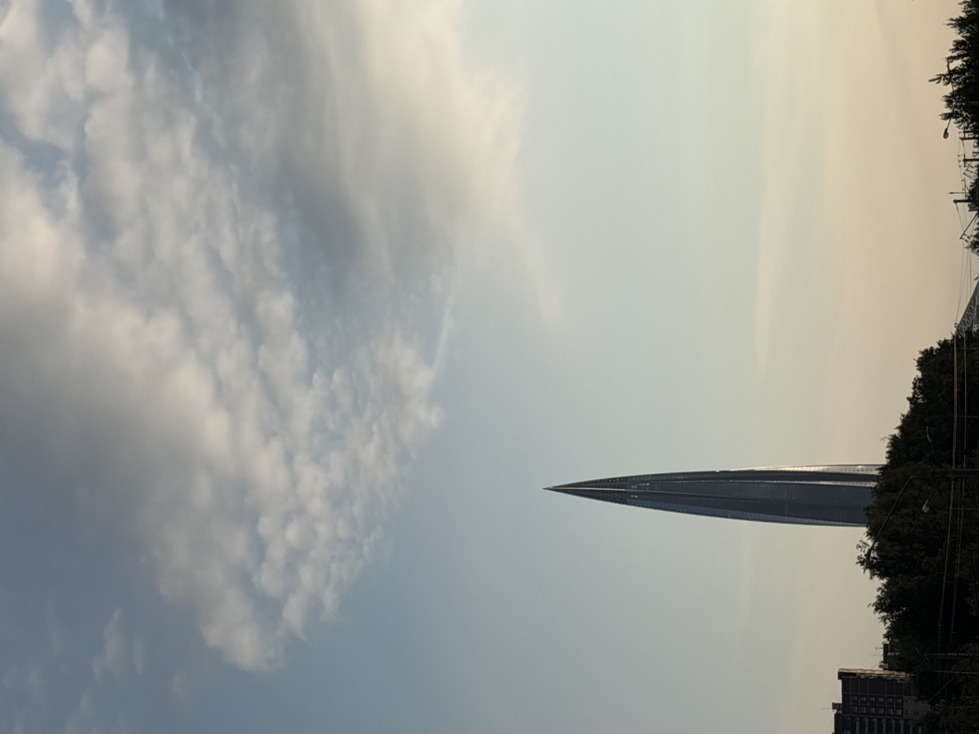 An exterior, medium shot captures the Lakhta Center skyscraper piercing the sky in St. Petersburg, Russia. The modern, glass-encased building is a dominant vertical element in the scene, its pointed spire reaching towards a sky filled with soft, diffused clouds. Below the skyscraper, a line of dark green trees and foliage forms a dense band across the lower portion of the frame, partially obscuring the base of the building and a few indistinct structures. A multi-story apartment building, with a reddish-brown brick facade, is visible to the left of the Lakhta Center, offering a contrast in architectural style. The overall lighting suggests either early morning or late afternoon, with a gentle, even illumination across the scene.